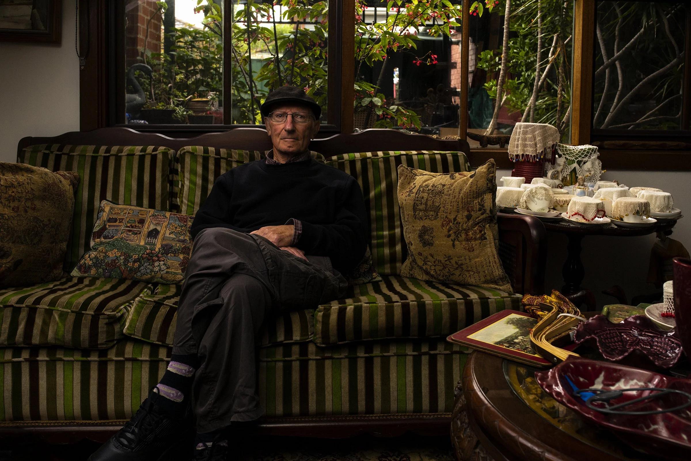 An elderly man wearing glasses, a black sweater, and a black cap sitting on a striped green and brown sofa next to decorative pillows, in a cozy room with large windows showing a lush backyard with plants and a decorative swan. There is a table with 