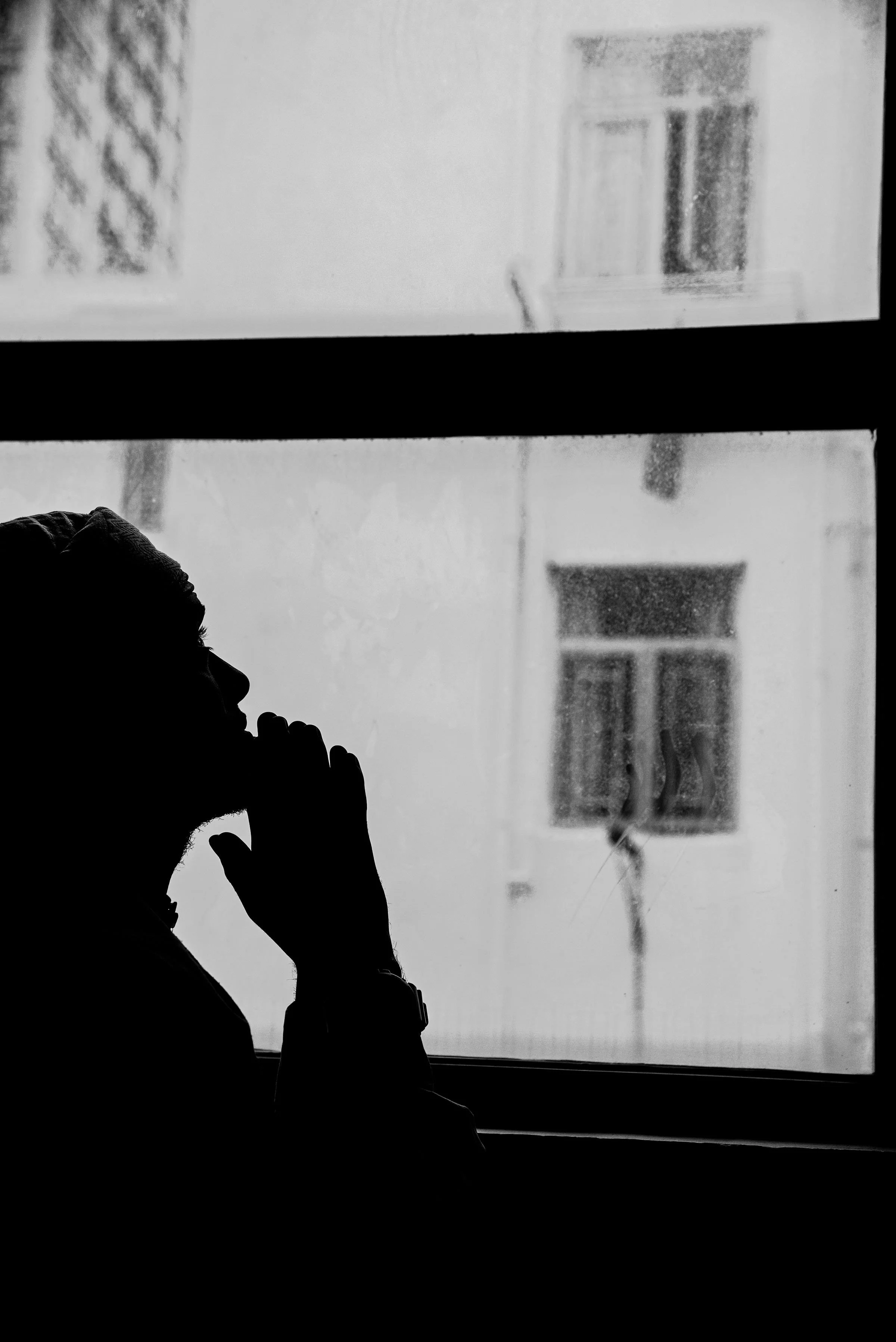 Silhouette of a person wearing a hoodie, talking on a cell phone, sitting near a window with raindrops and blurry buildings outside.