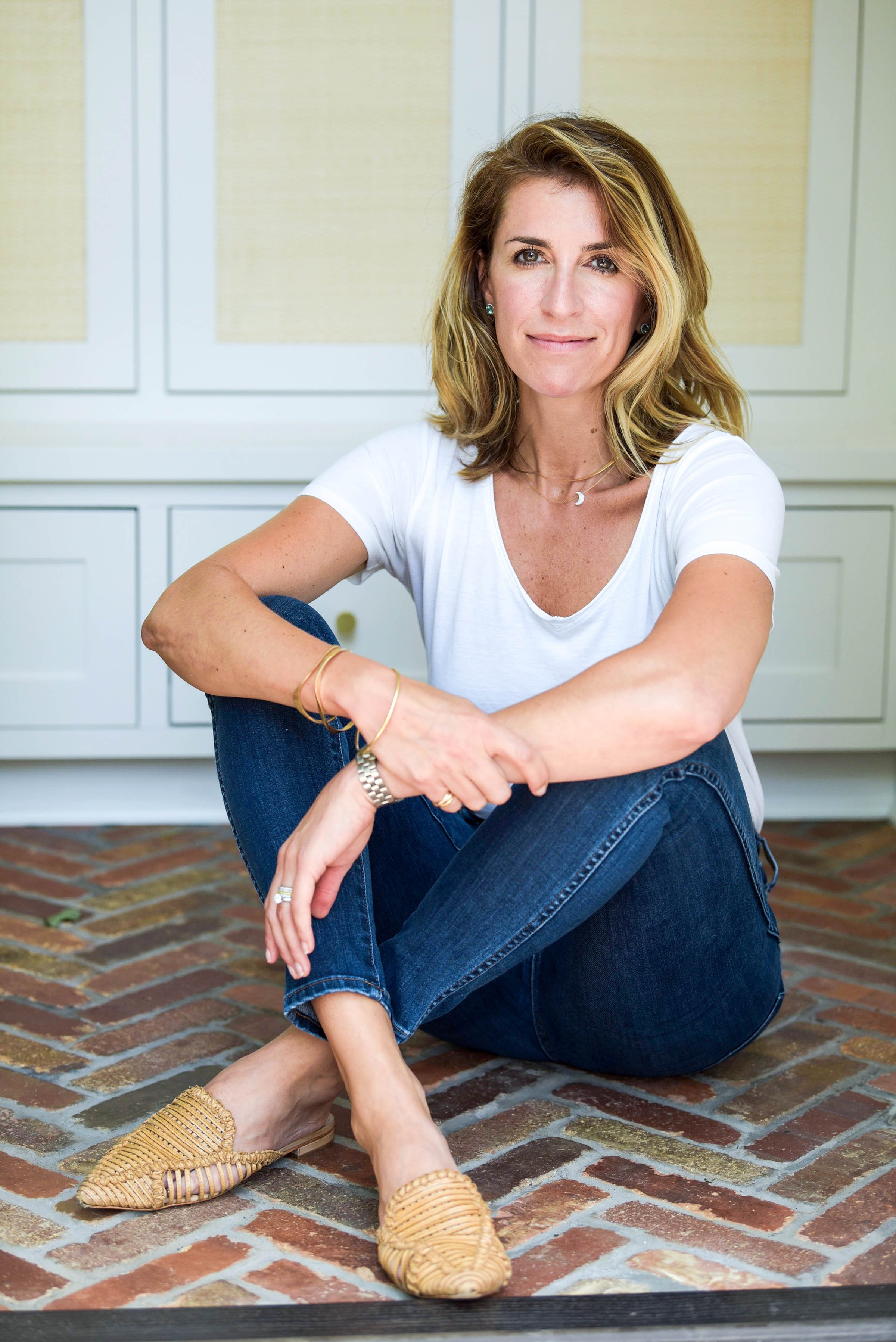 Portrait of a smiling woman with shoulder-length wavy blonde hair, wearing a white t-shirt, dark jeans, and tan woven shoes, sitting on a brick floor in front of white cabinets.