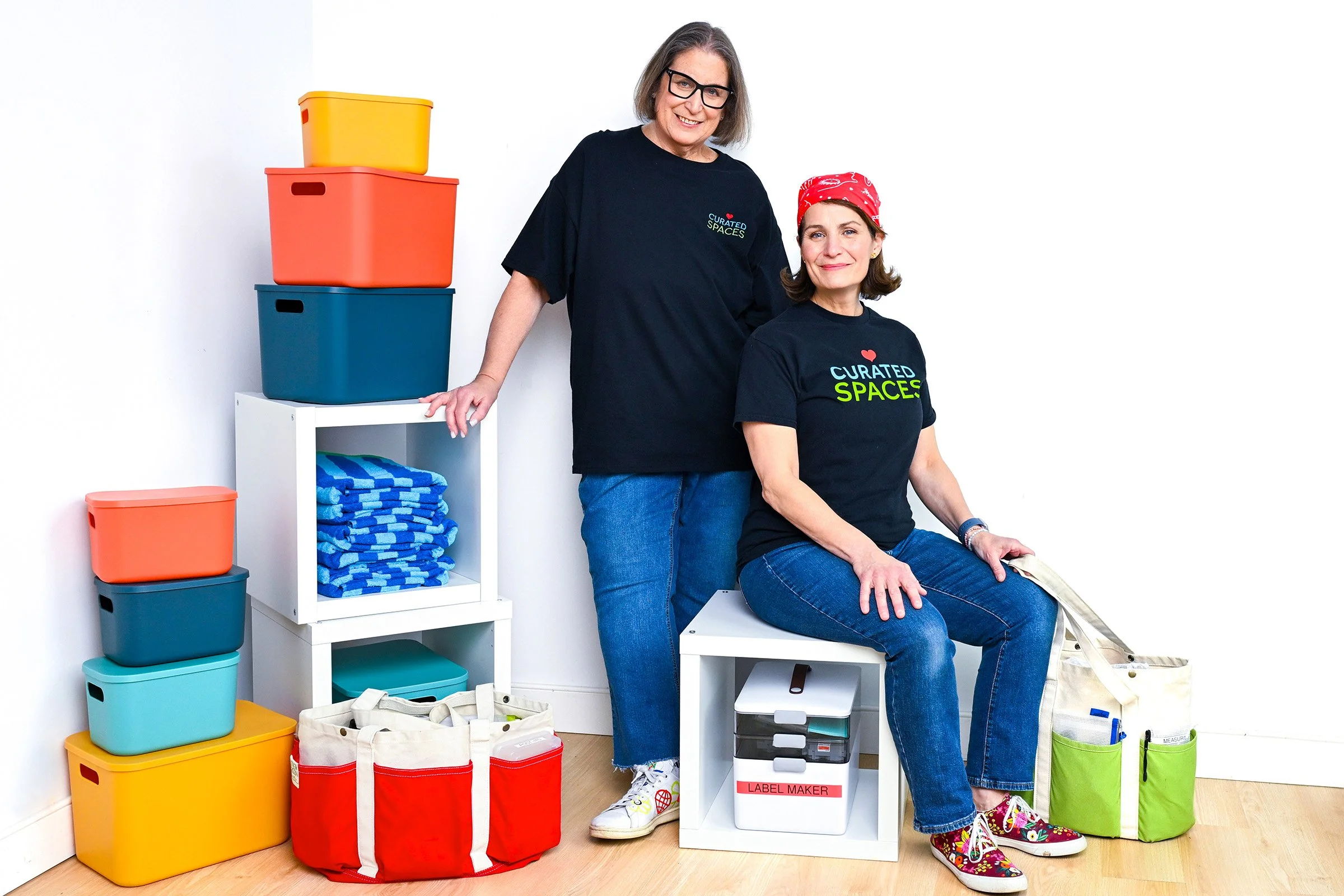 Two women in black T-shirts with 'CURATED SPACES' logo, one older with glasses and gray hair, the other younger with a red bandana, surrounded by colorful storage containers, shelves with folded towels, reusable grocery bags, and a label maker, in a 