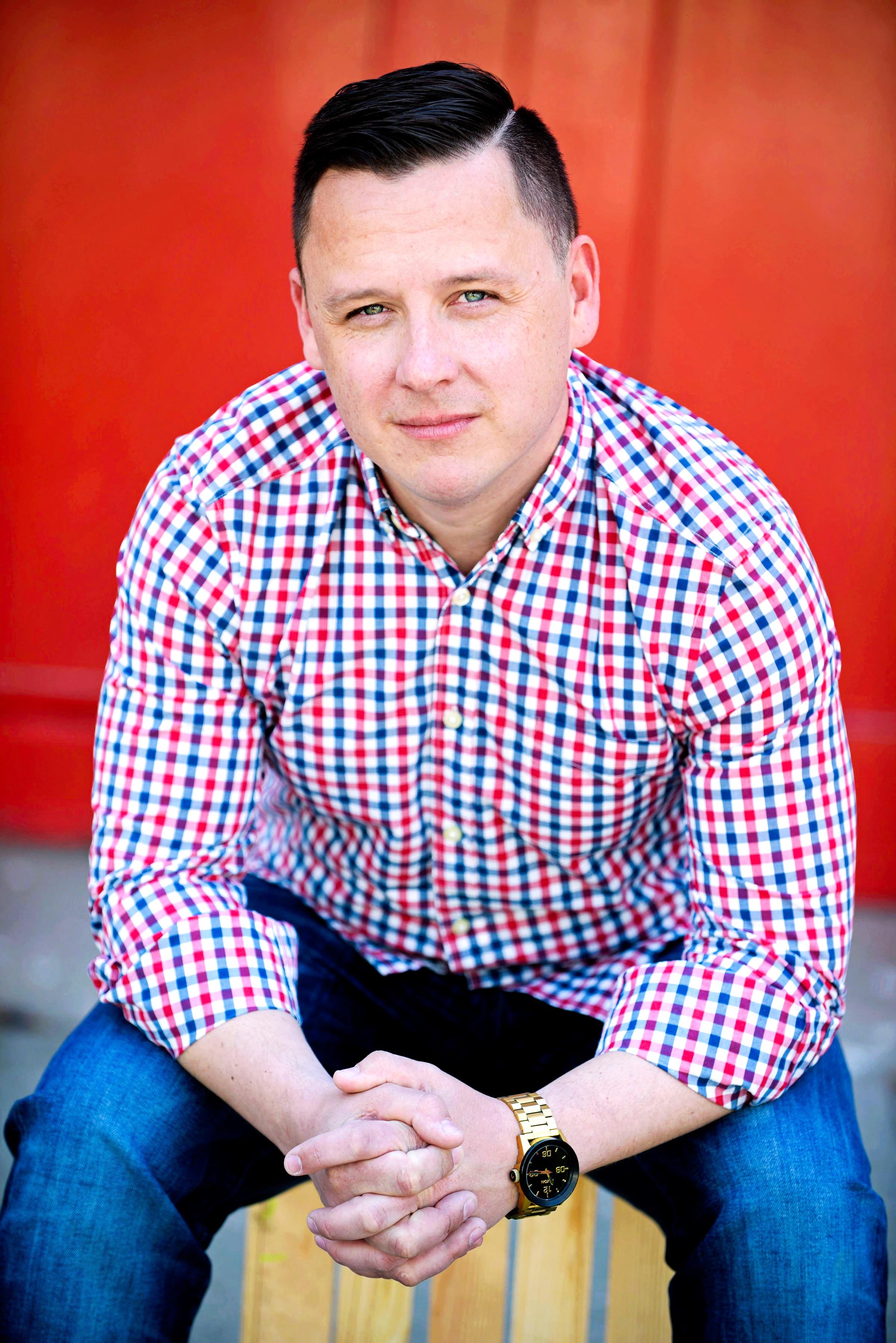 A man with short dark hair and fair skin wearing a red, white, and blue checkered shirt and blue jeans, sitting outdoors on a wooden chair against a red background, with his hands clasped and wearing a gold watch.