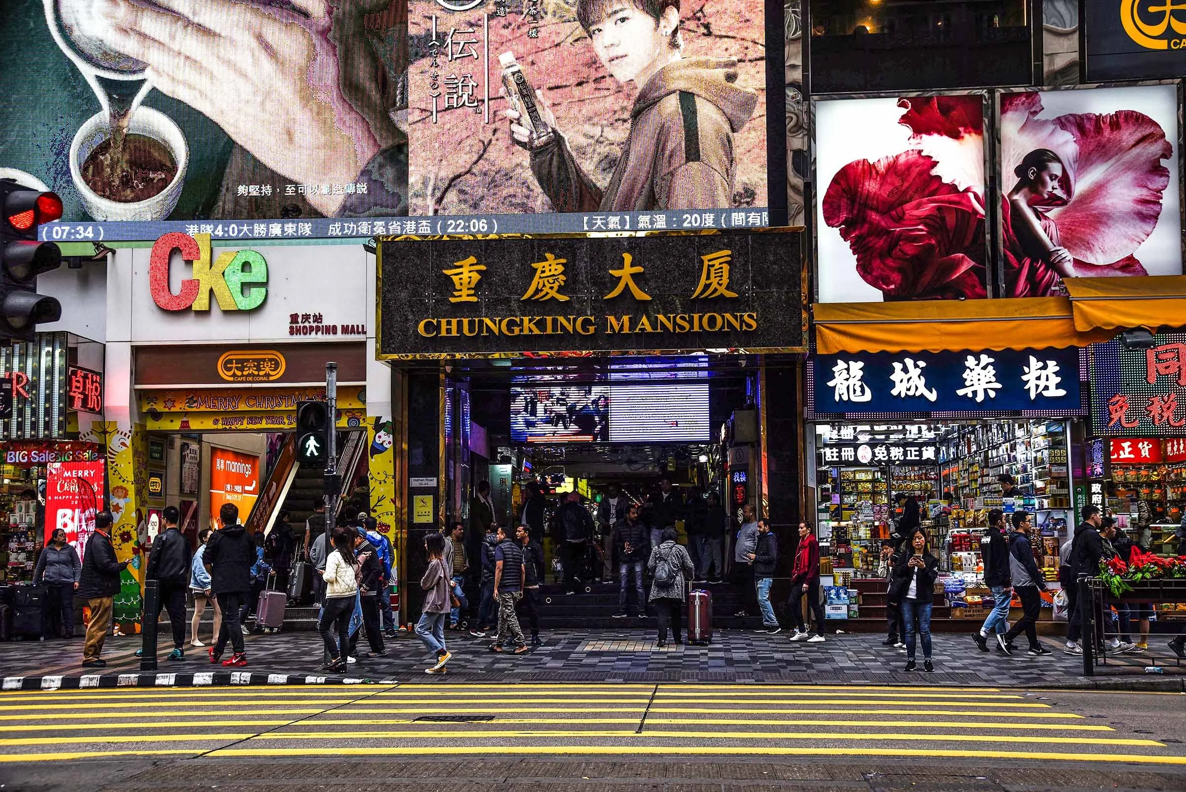 Crowded street scene in an urban shopping district with billboards and advertisements, including large digital screens displaying images and text, with people walking and standing on the sidewalk and a pedestrian crossing in the foreground.