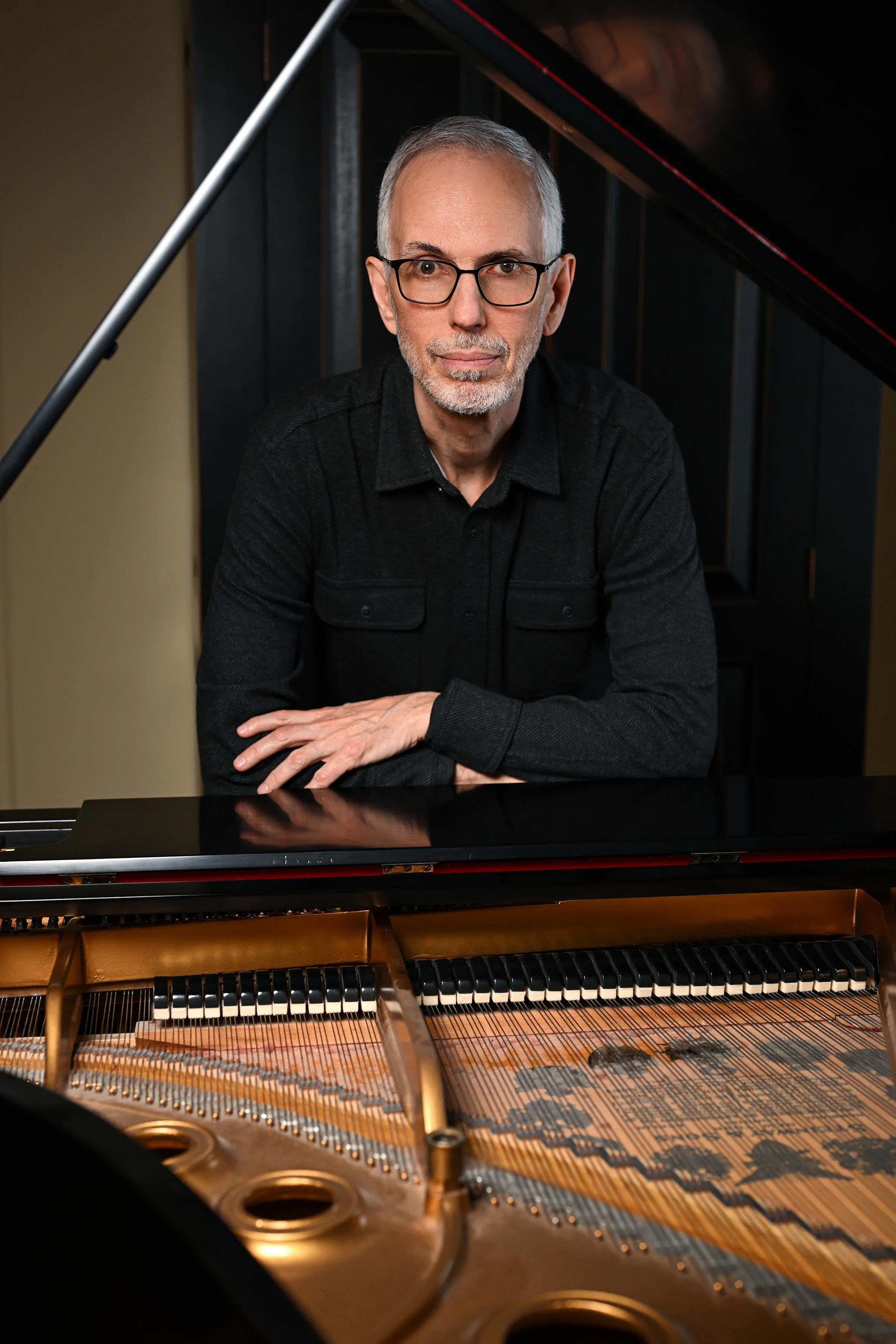 A man with gray hair, glasses, and a beard sitting at a grand piano with the lid open, looking directly at the camera.