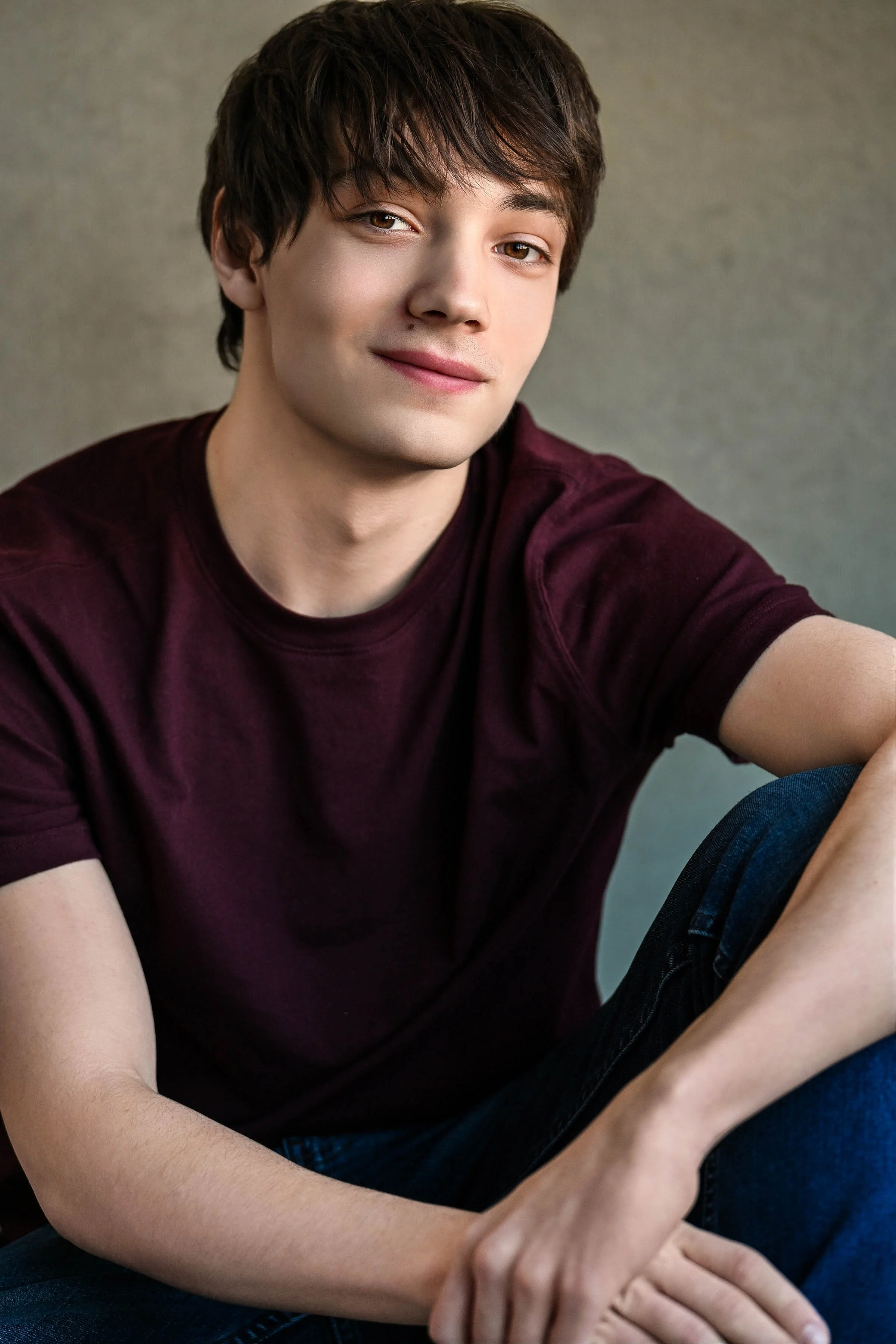 Close-up of a young man with brown hair, wearing a maroon T-shirt and sitting with his arms resting on his knee, smiling softly at the camera.