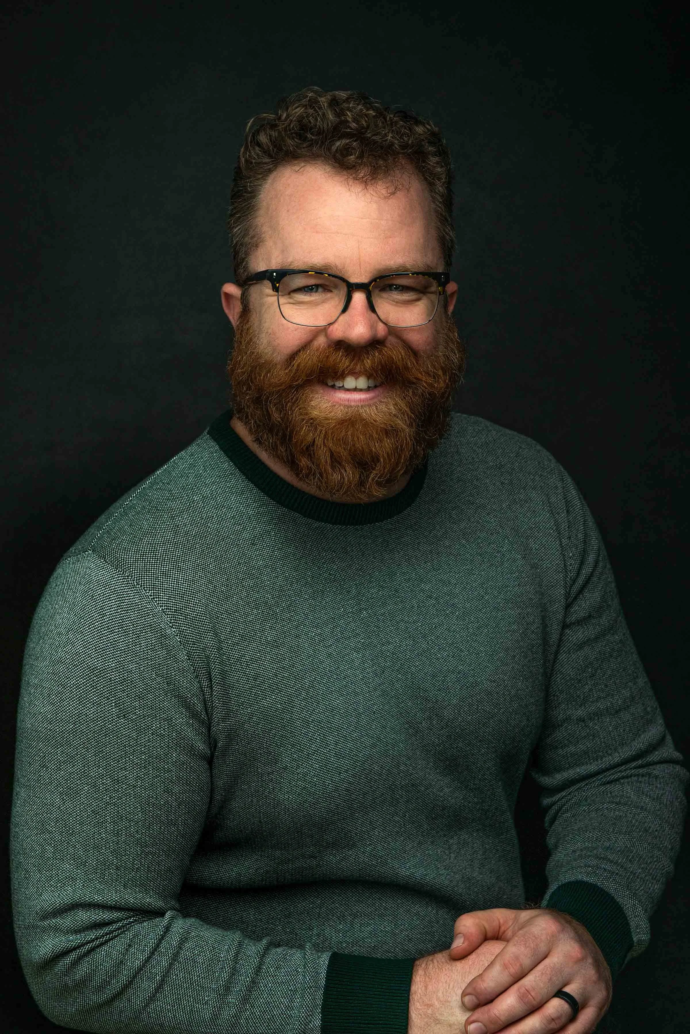 Close-up of a smiling man with curly hair, glasses, a beard, and wearing a gray sweater with black accents, against a dark background.
