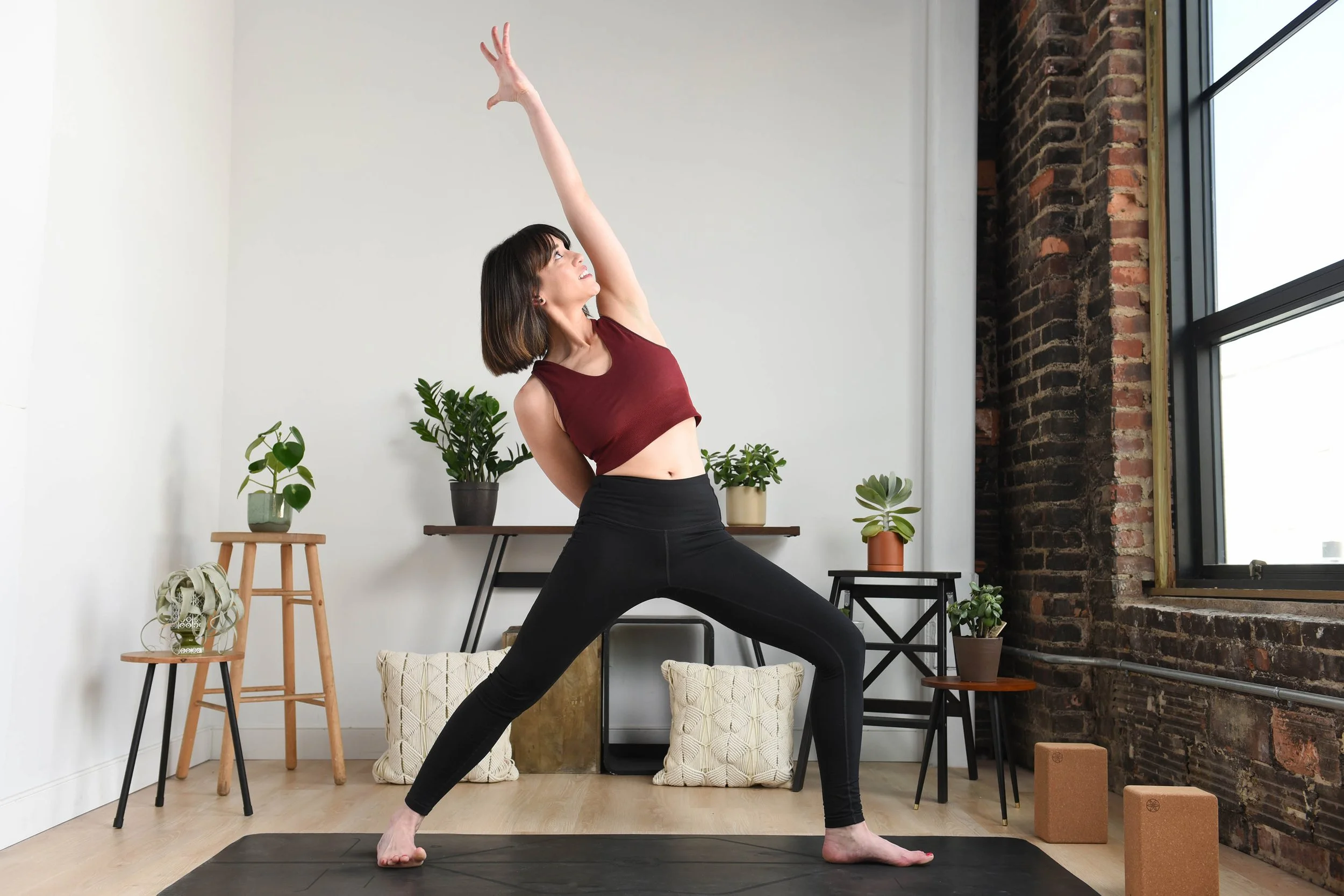 A woman practicing yoga in a studio with plants and brick wall, wearing a red tank top and black leggings.