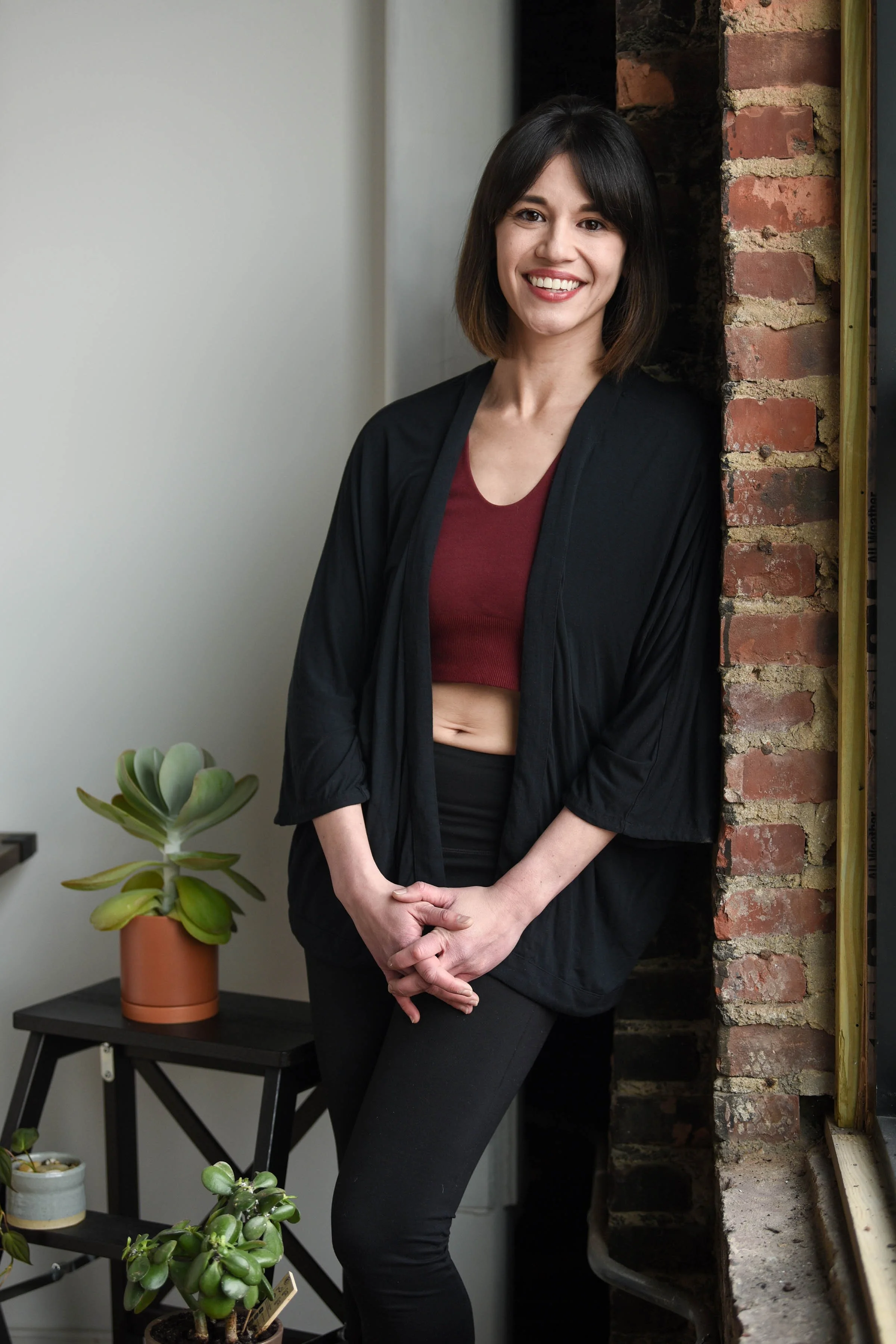 A woman with shoulder-length dark hair smiling and standing near a brick wall indoors. She is wearing a black jacket over a red crop top and black pants. There are potted plants on a black table nearby.