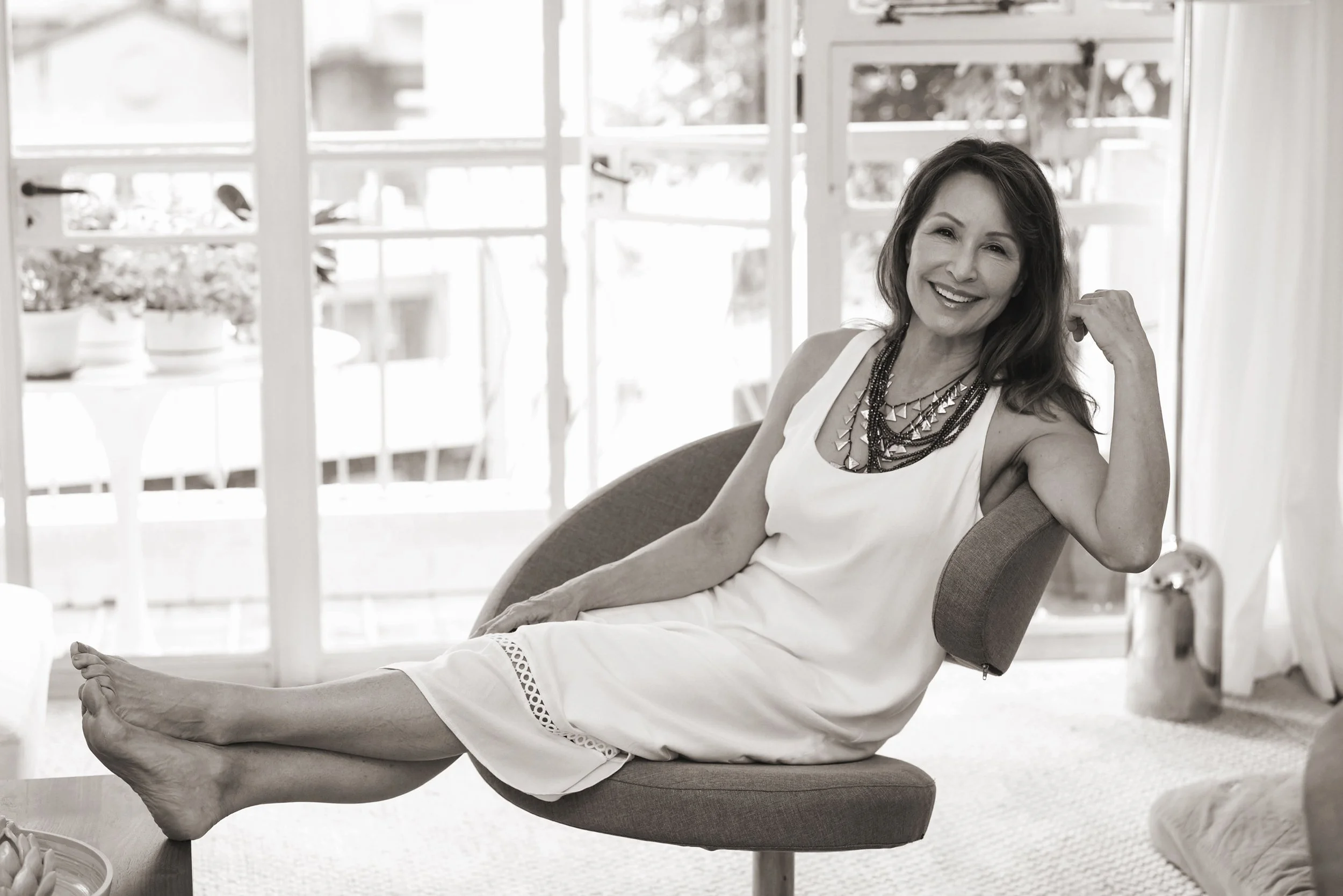 A woman with a big smile, wearing a white dress and multiple layered necklaces, sitting cross-legged on an office chair and relaxed in a bright room with large windows.