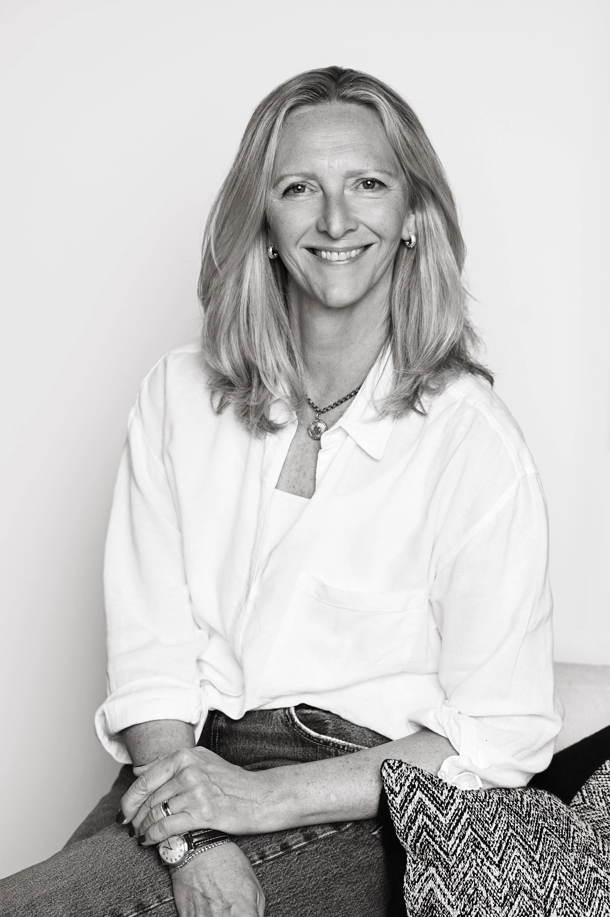 Black and white portrait of a smiling woman with shoulder-length hair, wearing earrings, a necklace, a button-up shirt, and a watch, sitting with her hands crossed.