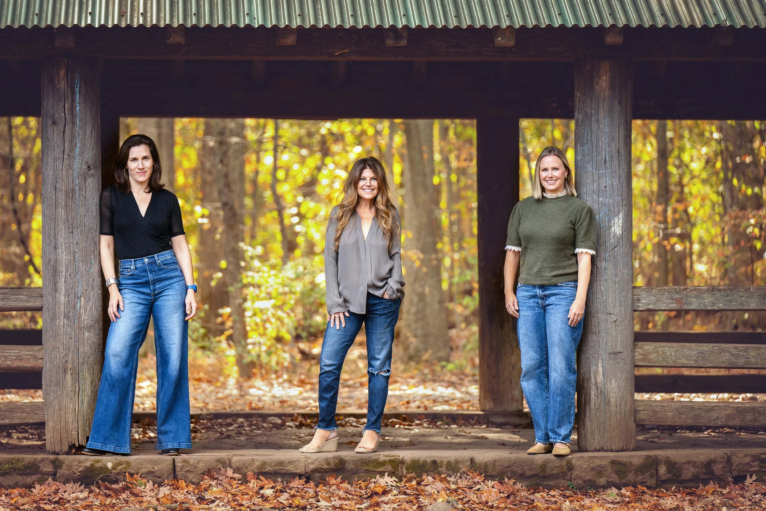 Three women standing outdoors on a wooden bridge in a forest during autumn, with colorful fallen leaves on the ground, smiling and looking at the camera.