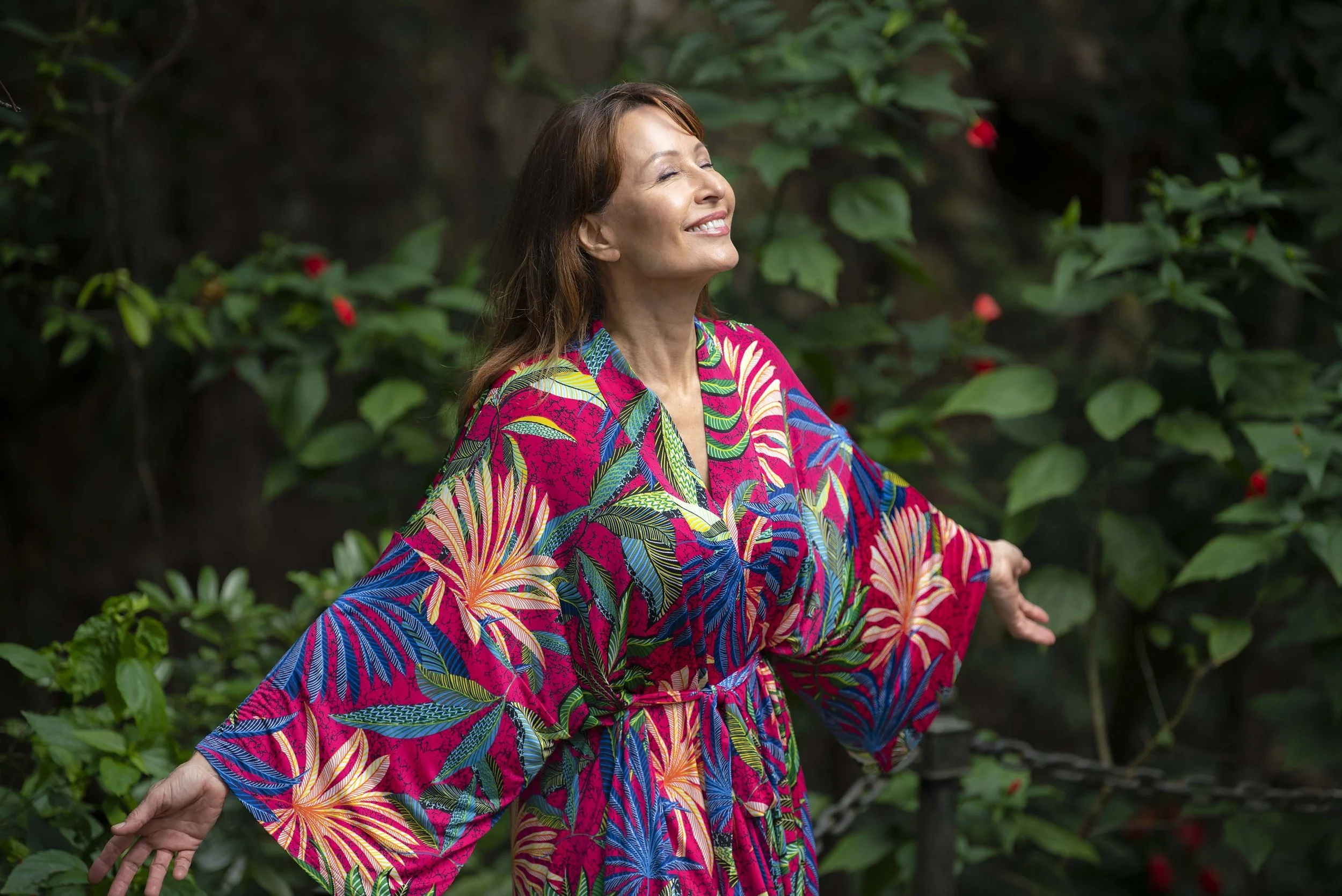 A woman with closed eyes and a smile, wearing a colorful, floral-patterned dress, standing outdoors among green plants with red flowers, her arms outstretched.