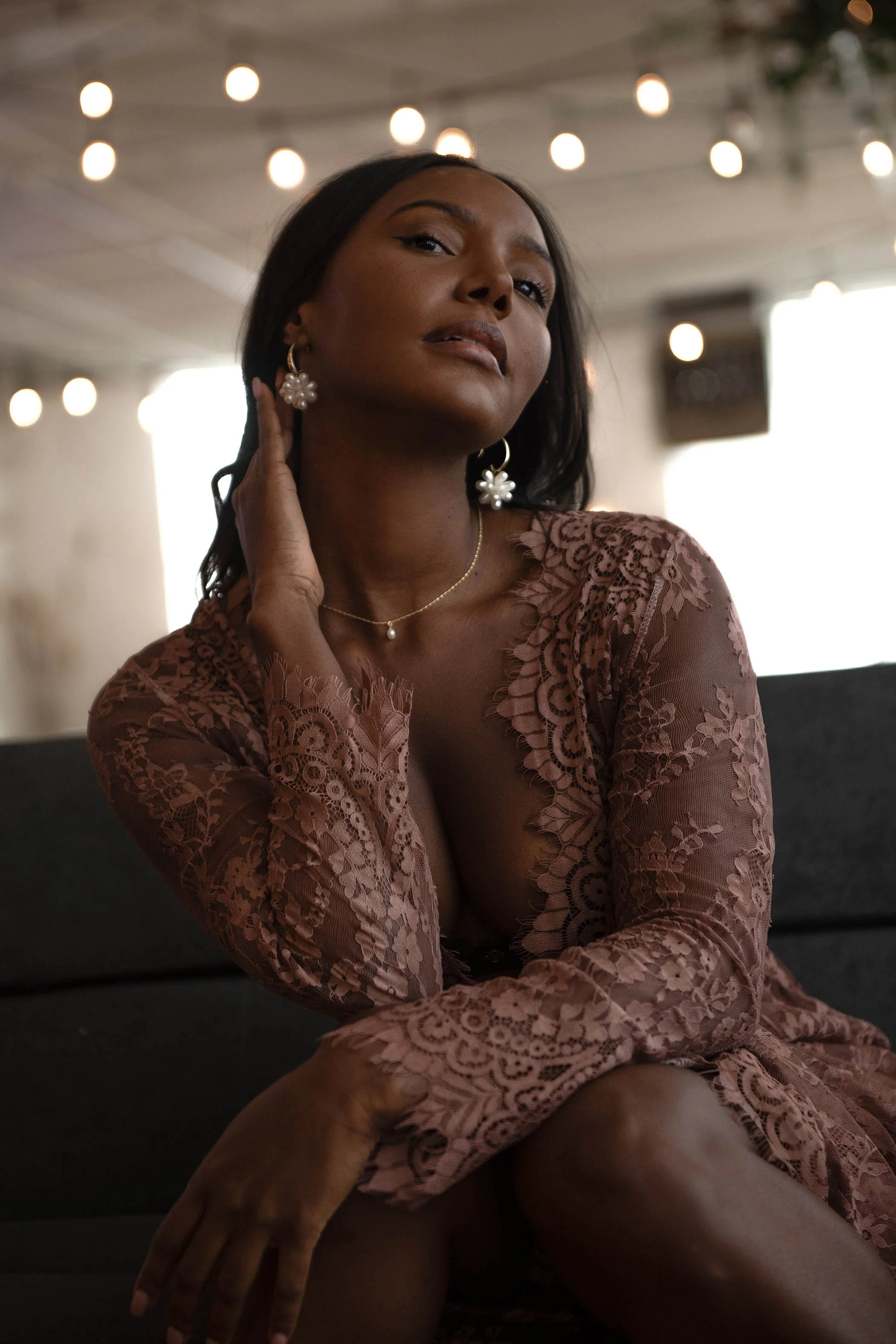 A woman with dark hair and medium skin tone poses in a lace dress with floral earrings and a pearl necklace, sitting on a dark surface with blurred lights in the background.