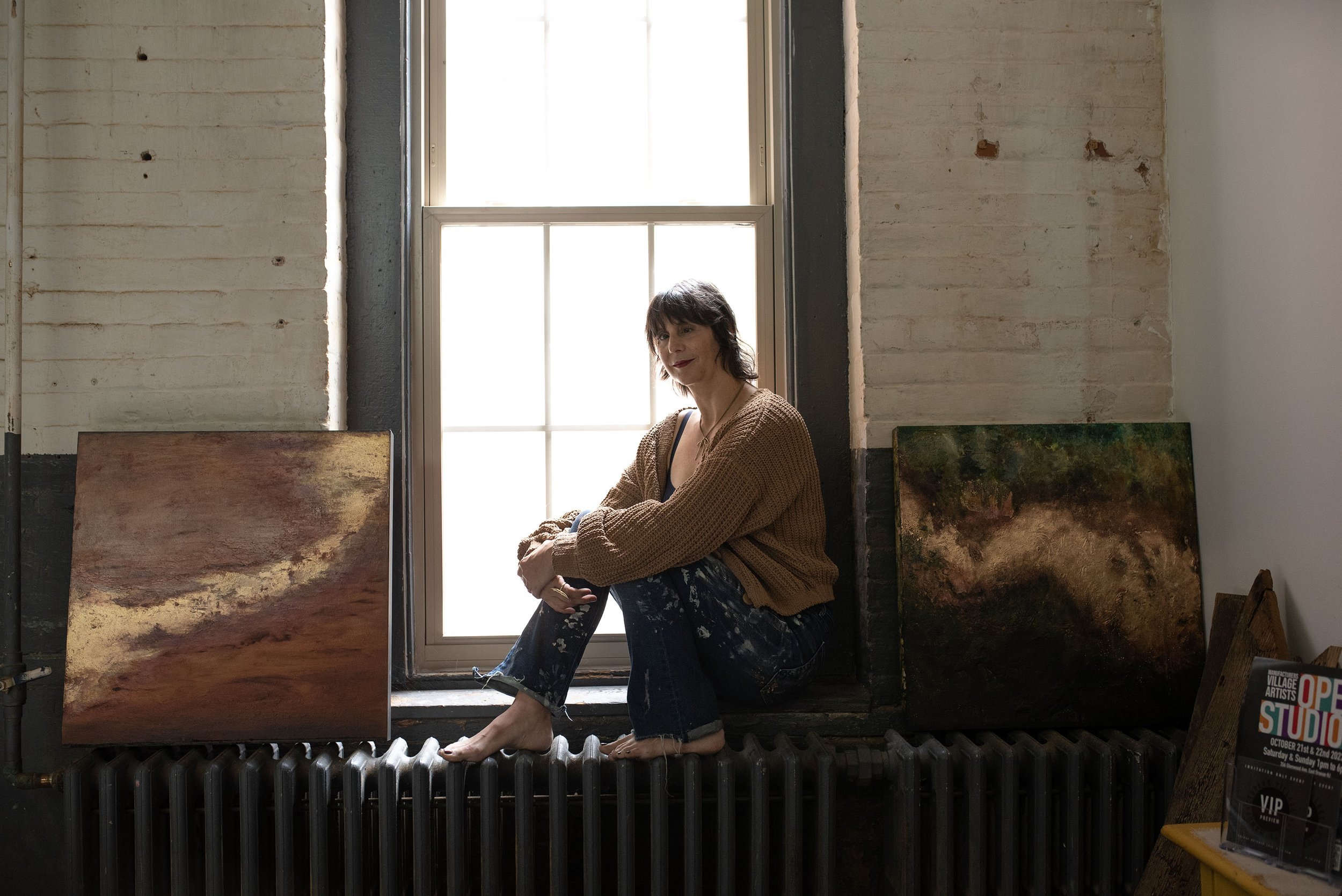 A woman with short dark hair and a brown sweater poses on a radiator in an art studio with paintings leaning against the wall behind her, a window providing natural light, and art promotional materials on a table in the corner.