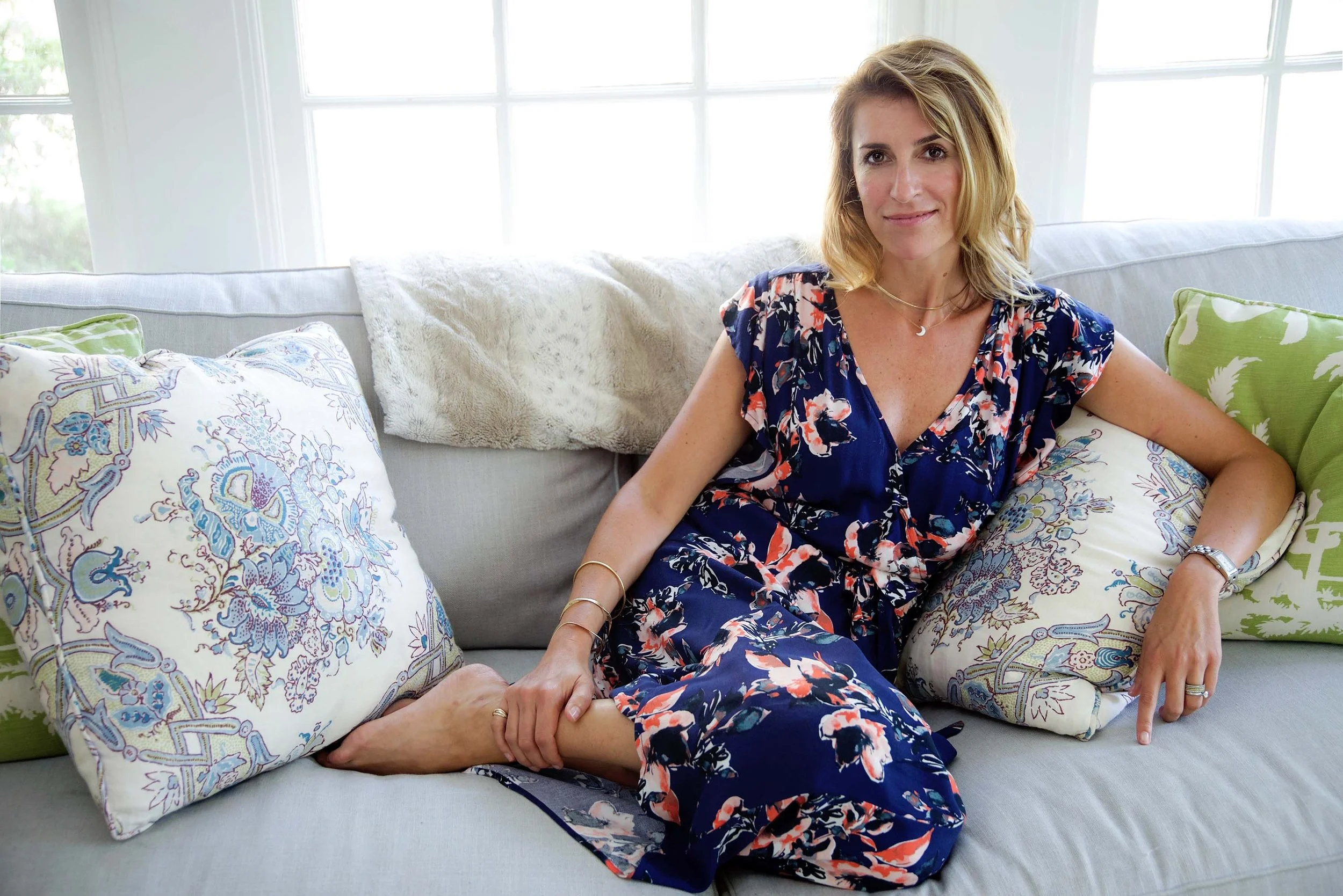 Woman sitting on a light-colored couch, smiling, with colorful pillows behind her. Bright windows in the background.