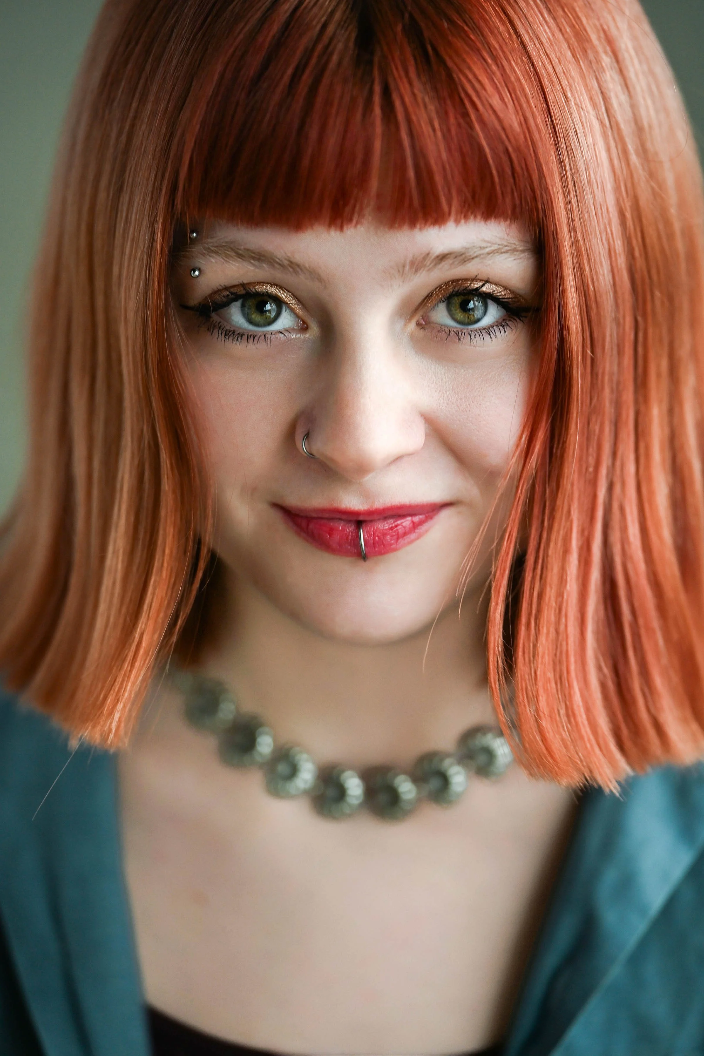 Close-up of a young woman with red hair, green eyes, gold eyeshadow, and piercings near her eyebrows and lower lip. She is wearing a pearl necklace and a teal top.