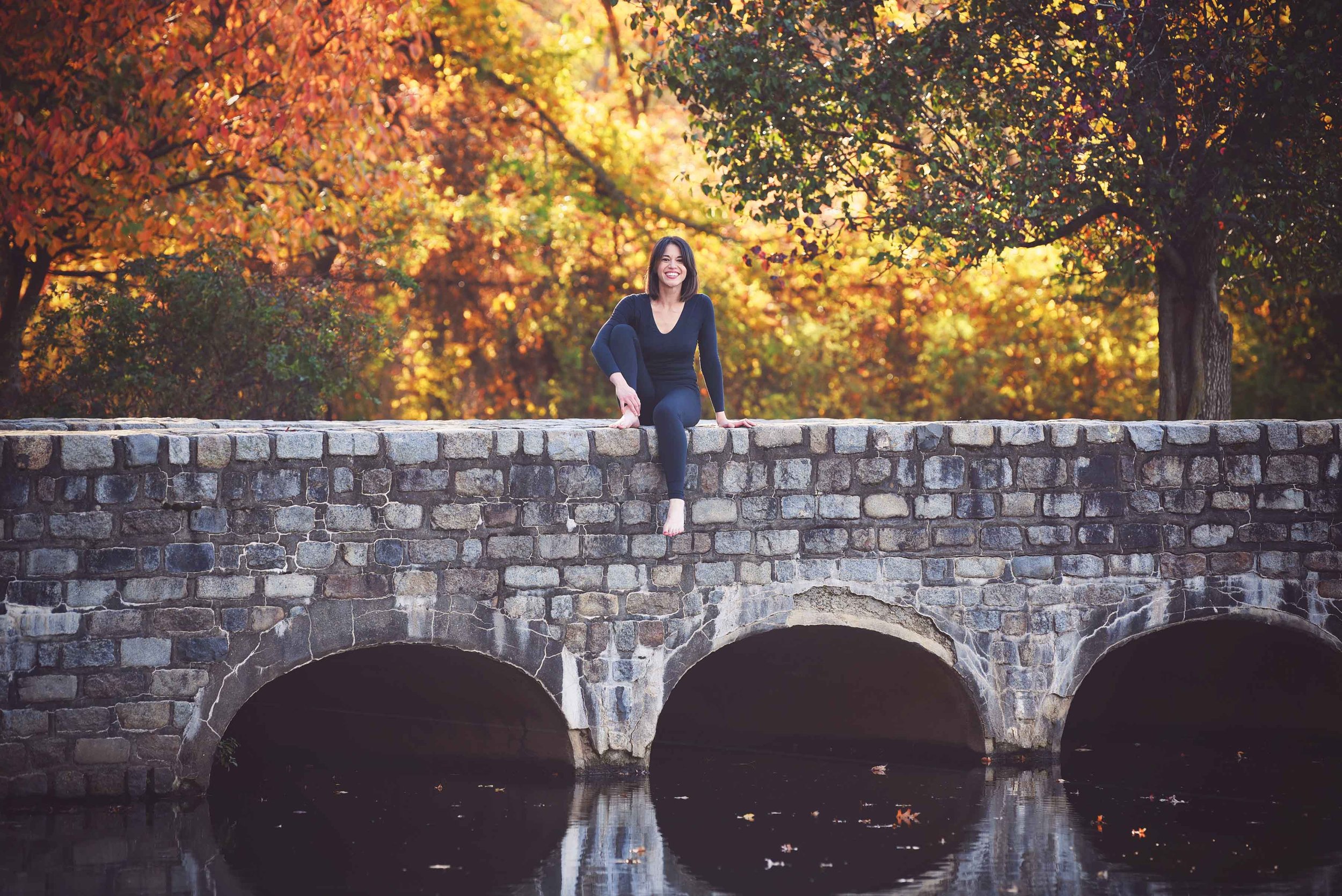 A woman in black clothing sitting on a stone bridge over a river with autumn foliage in the background.