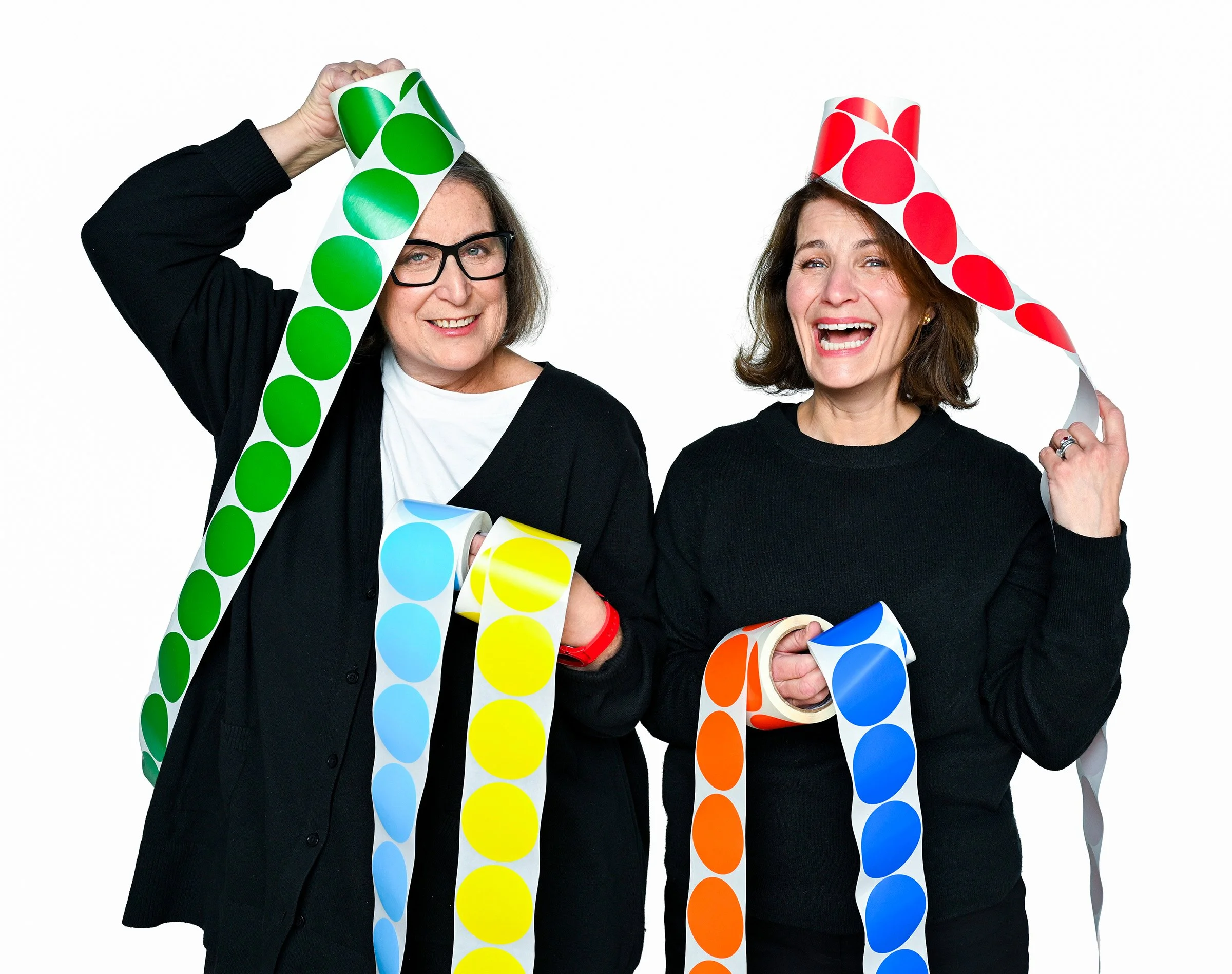 Two women smiling and holding colorful polka-dot paper rolls, one wearing glasses and the other with brown hair.