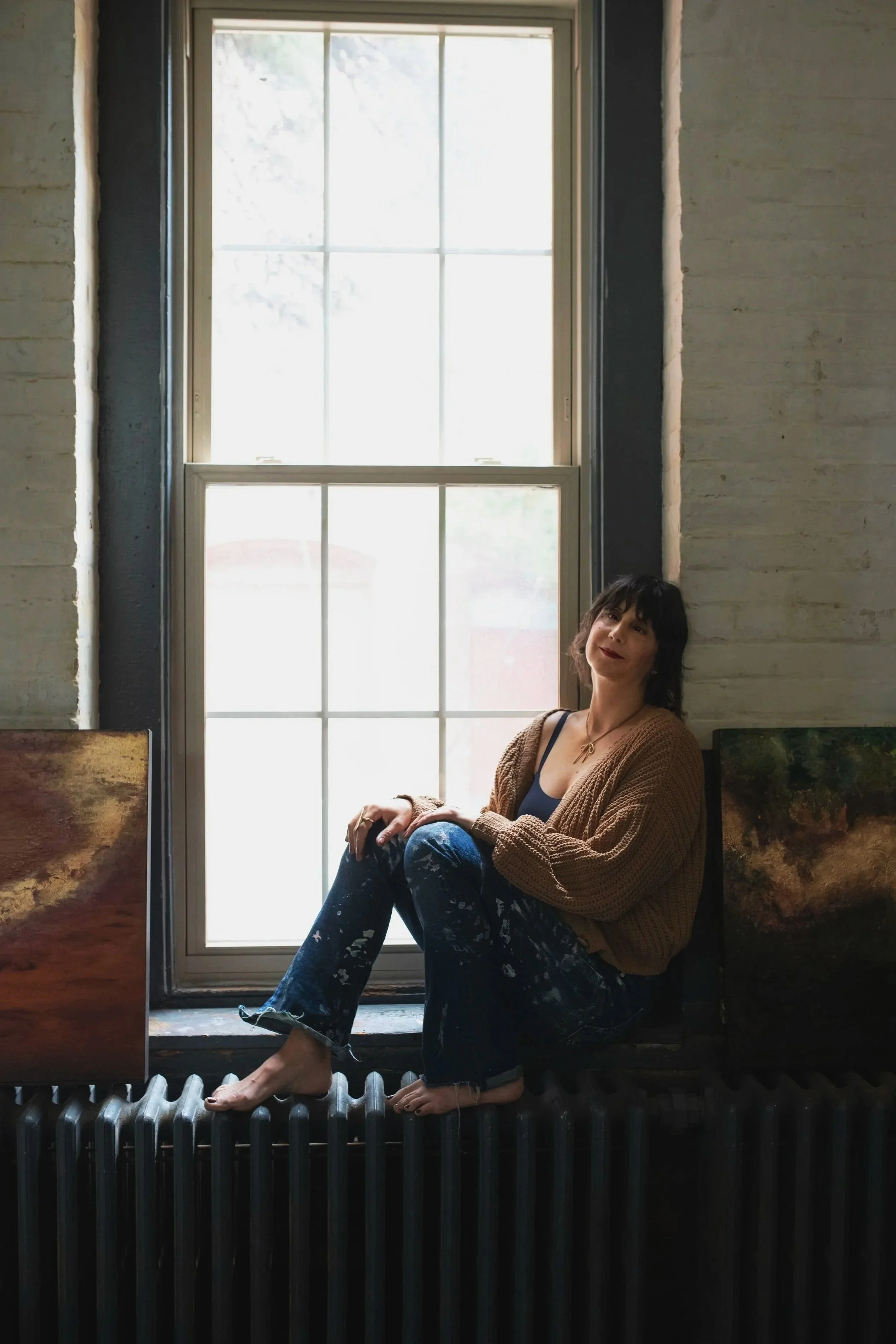 Woman sitting on a radiator in front of a large window, with paintings on either side, inside a room with brick walls.