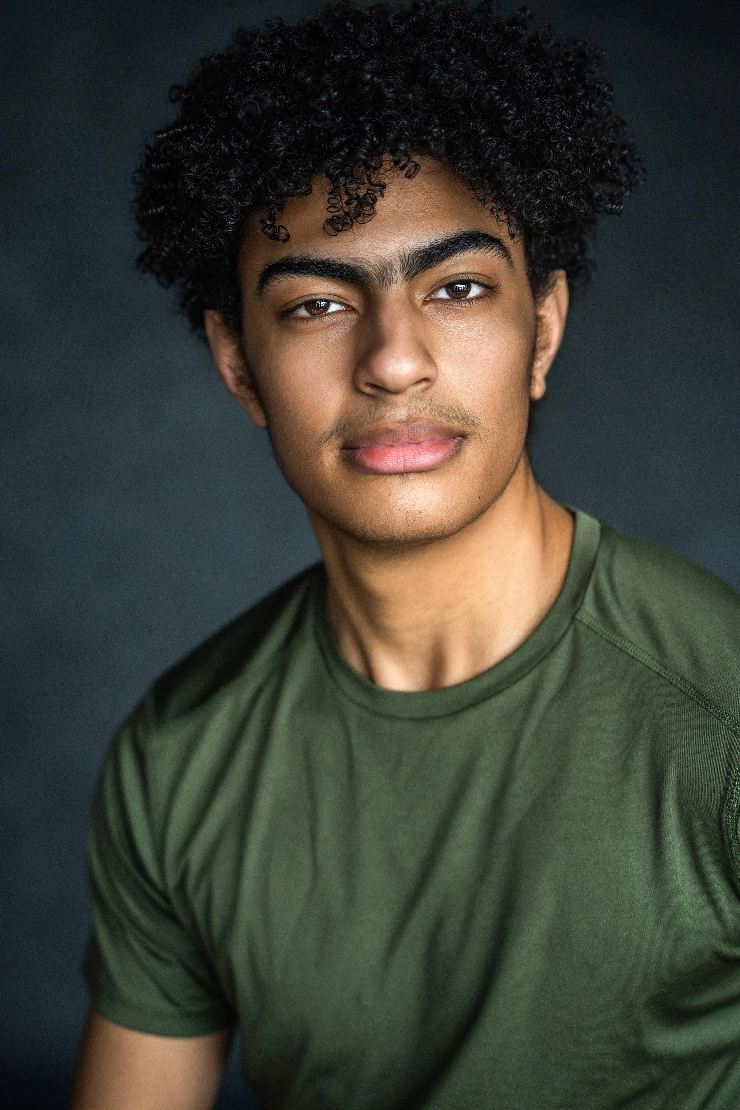 Close-up of a young man with dark curly hair, light brown skin, and wearing a green shirt, looking slightly to the right with a neutral expression, against a dark, plain background.
