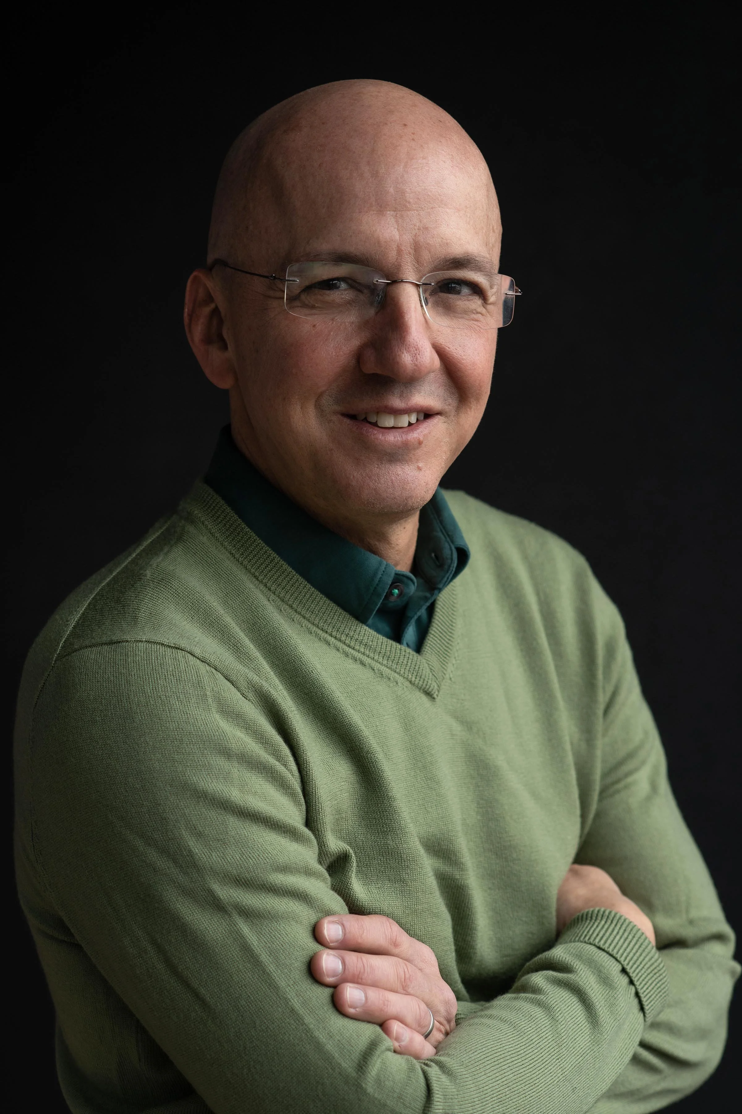 A middle-aged man with a bald head and glasses is smiling at the camera. He is wearing a green sweater over a dark shirt and has his arms crossed in front of him. The background is black.