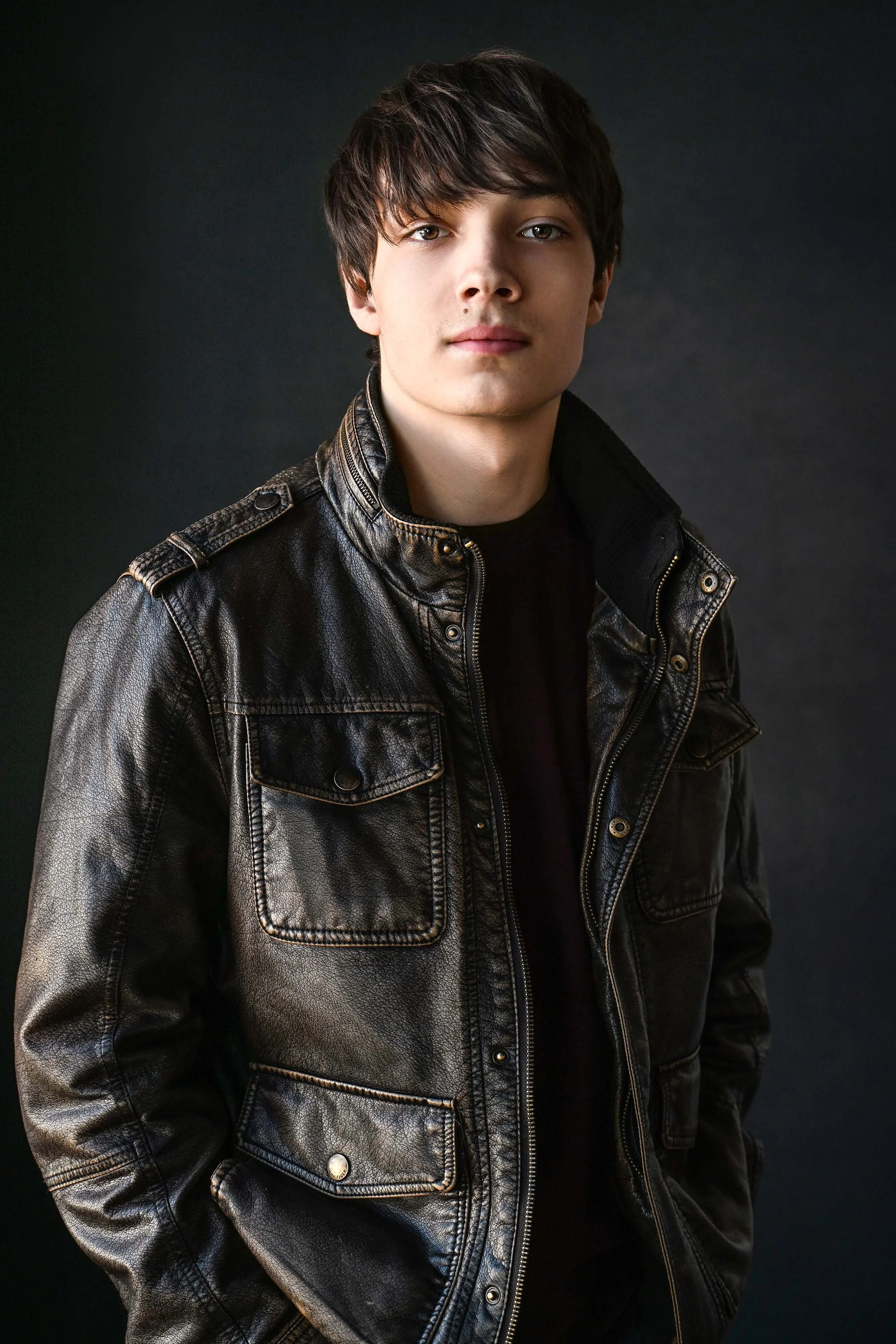 A young man with brown hair wearing a black leather jacket against a dark background.