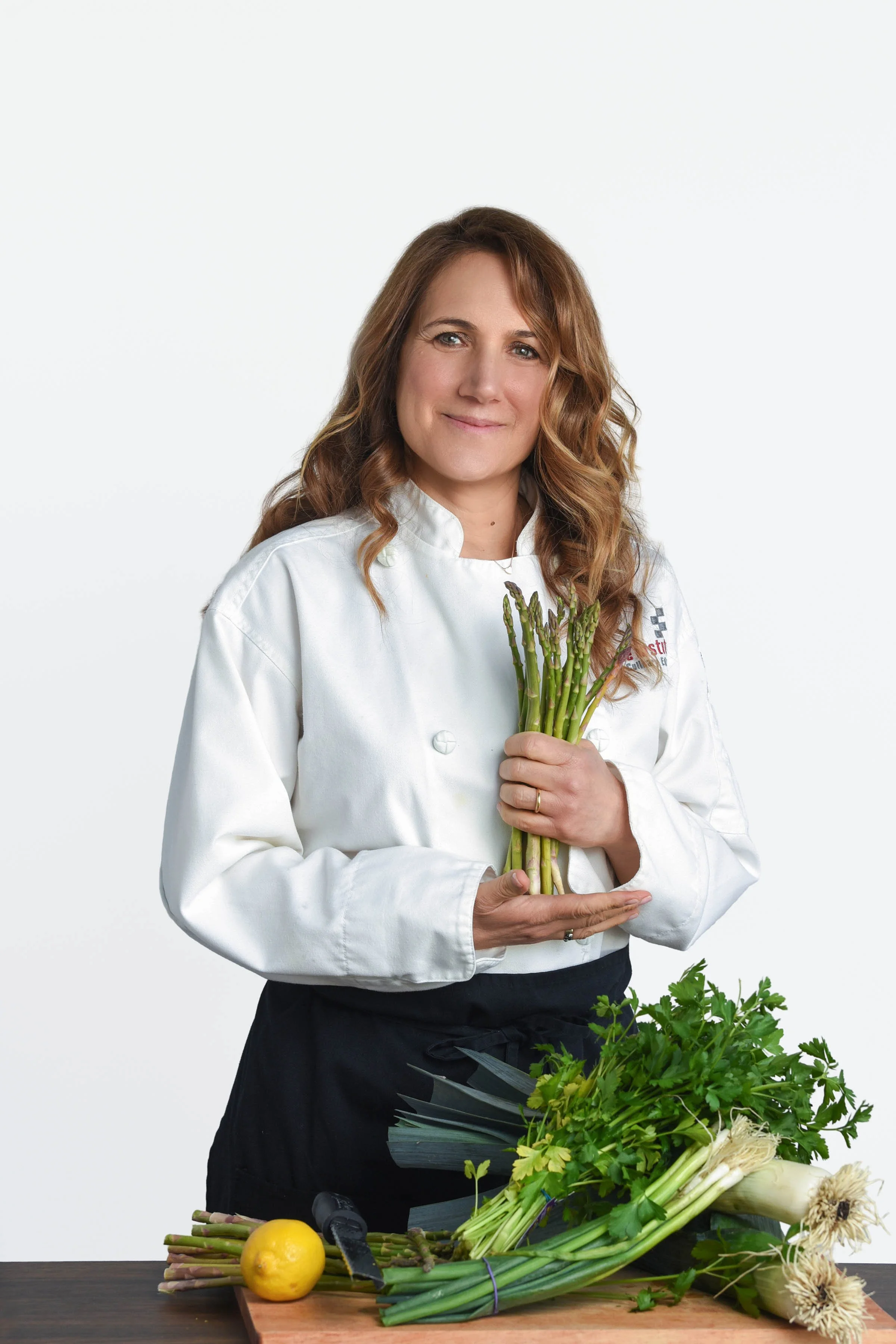 A woman in a white chef's coat holding a bunch of fresh asparagus. A cutting board with a lemon, more asparagus, and flowers is on the table in front of her, with green leafy herbs nearby.