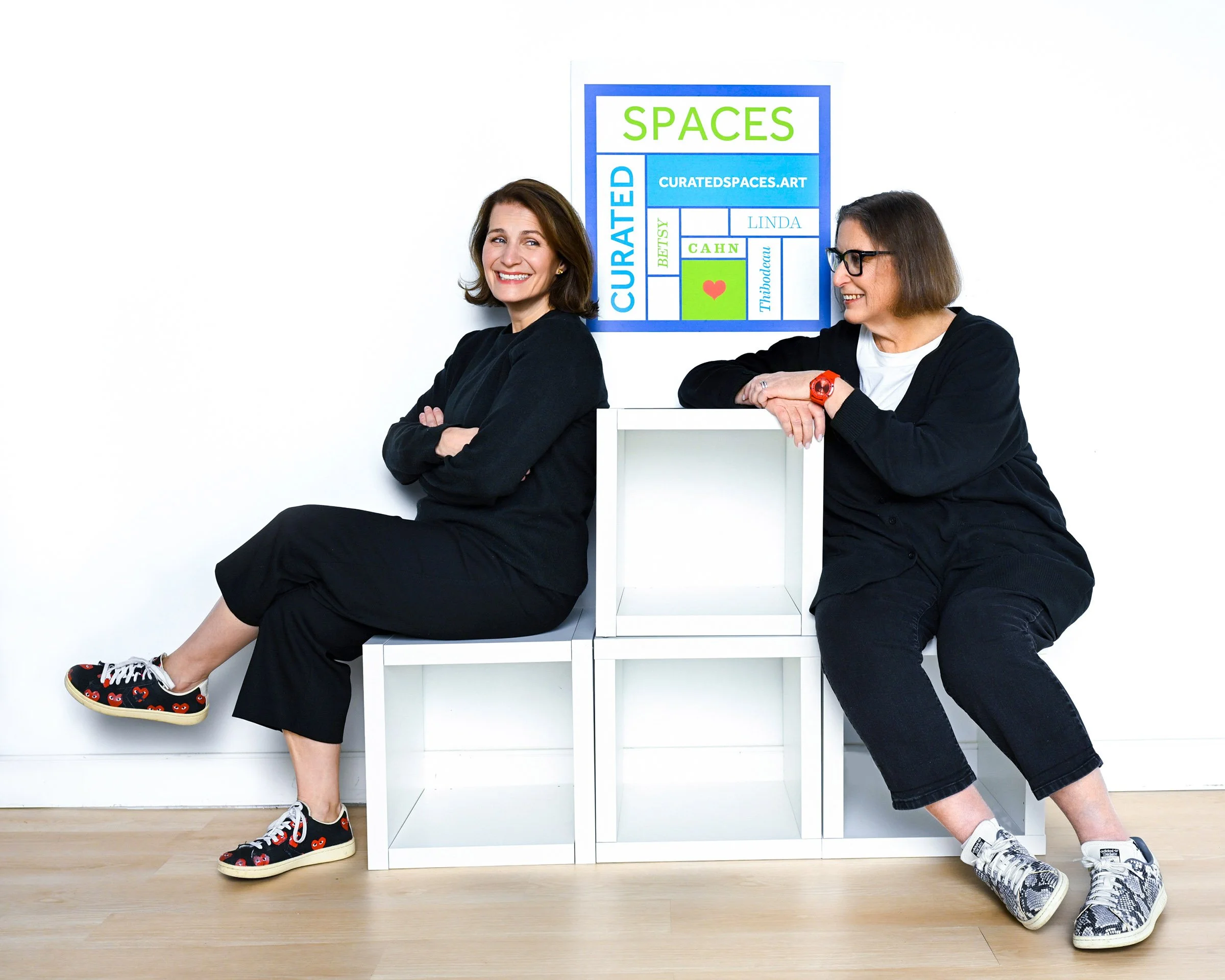 Two women sitting on a white shelf in front of a white wall with colorful art that says 'SPACES' and various words related to curated spaces. One woman is smiling with arms crossed, wearing black clothing and heart-themed sneakers. The other woman is