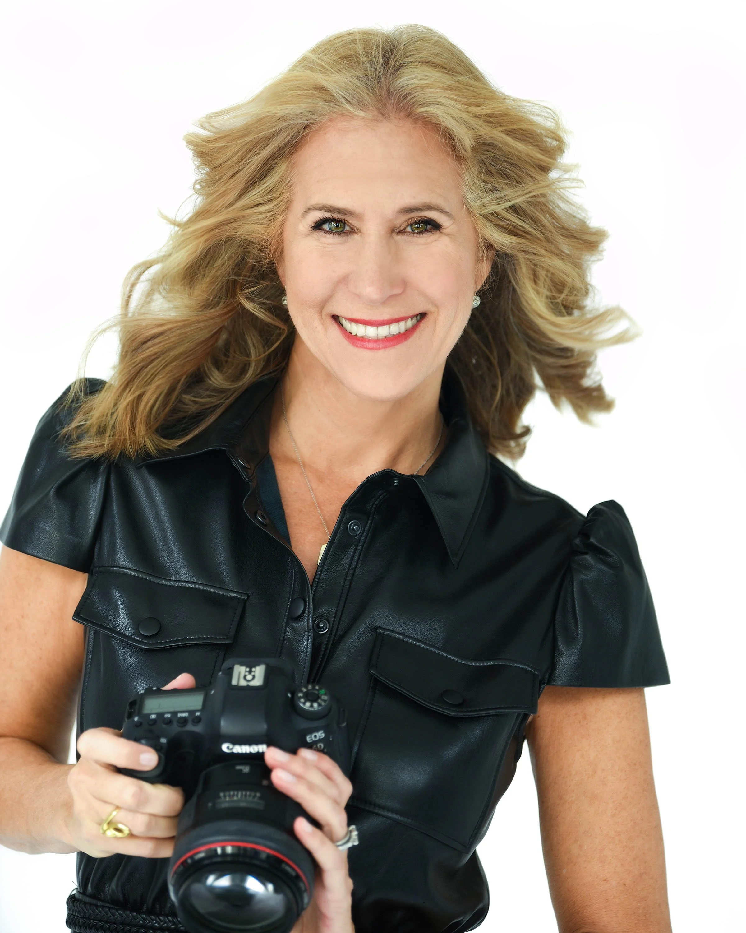 A woman with blonde hair, wearing a leather shirt, holding a Canon DSLR camera, smiling at the camera.