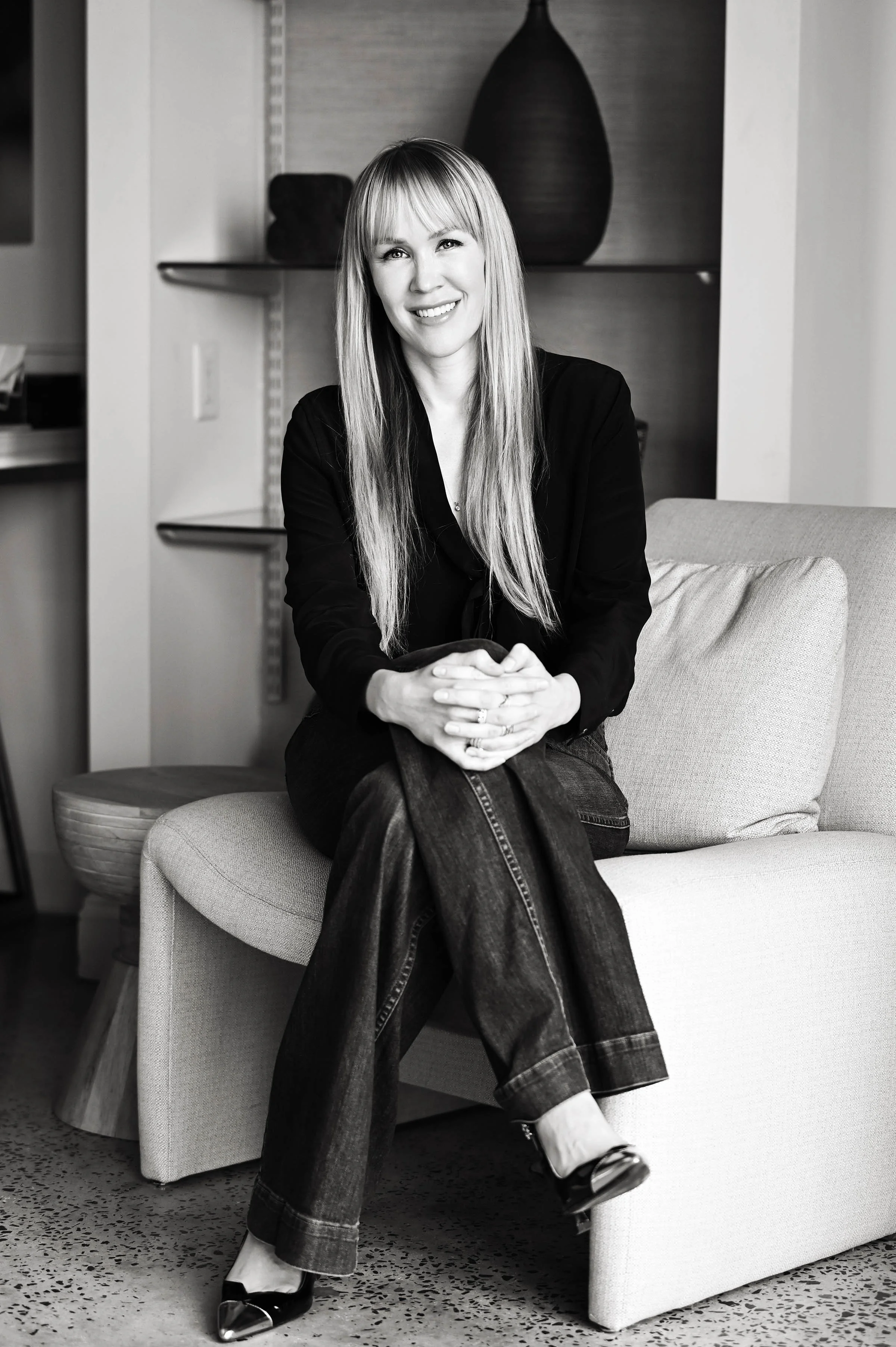 A woman with long hair sitting on a couch, smiling, with legs crossed, wearing a black jacket, jeans, and high heel shoes, in a modern interior setting.