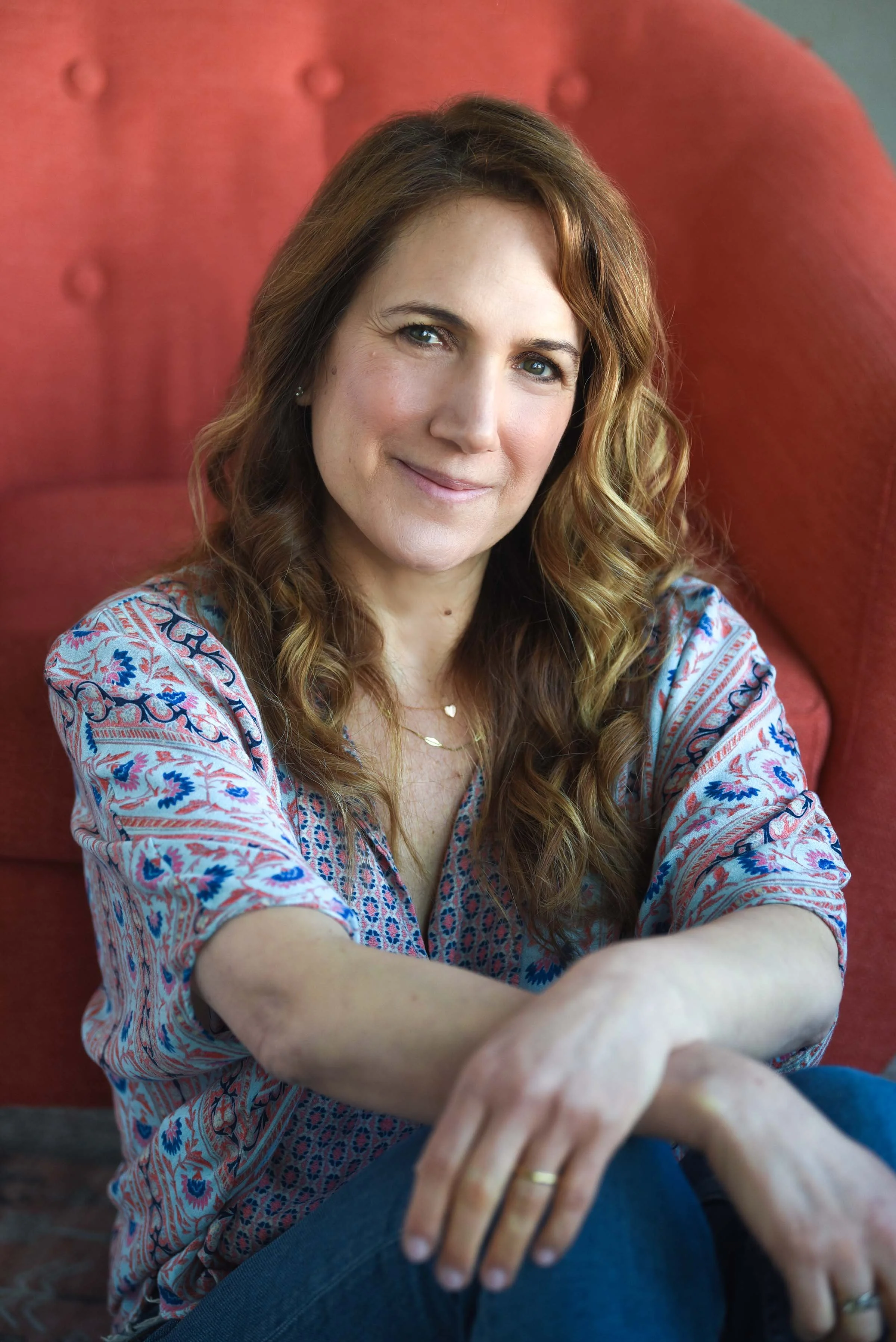 A woman with wavy brown hair and blue eyes smiling, wearing a patterned blouse, sitting on a red cushioned chair.