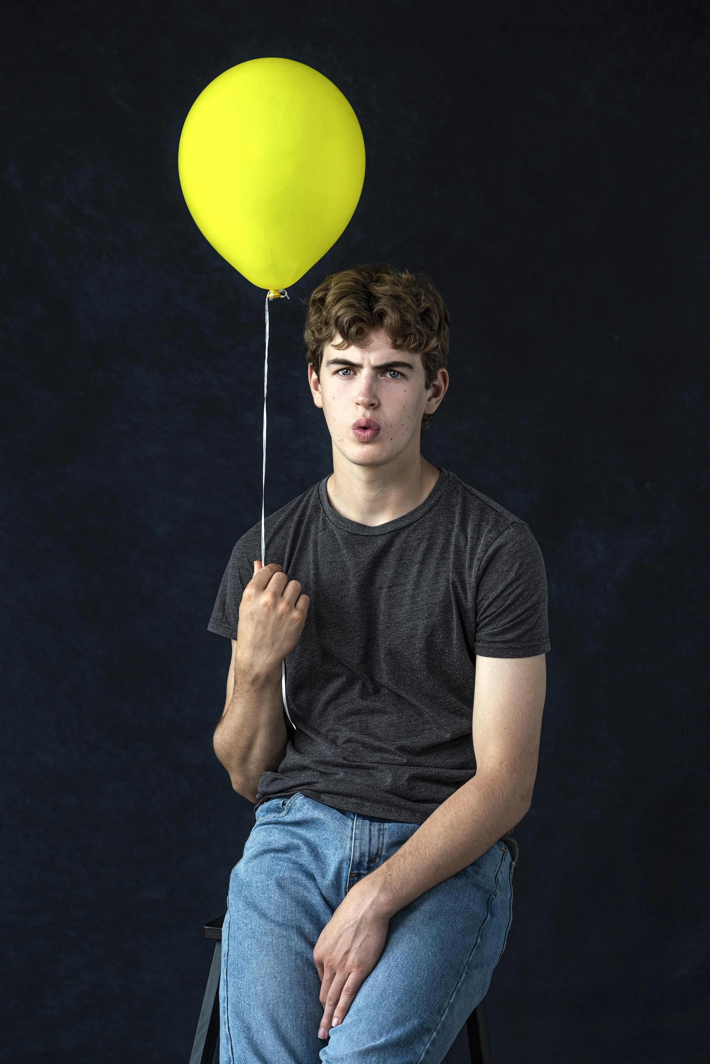 A young man with curly brown hair wearing a dark gray t-shirt and blue jeans sitting on a black stool against a dark textured background while holding a yellow balloon.