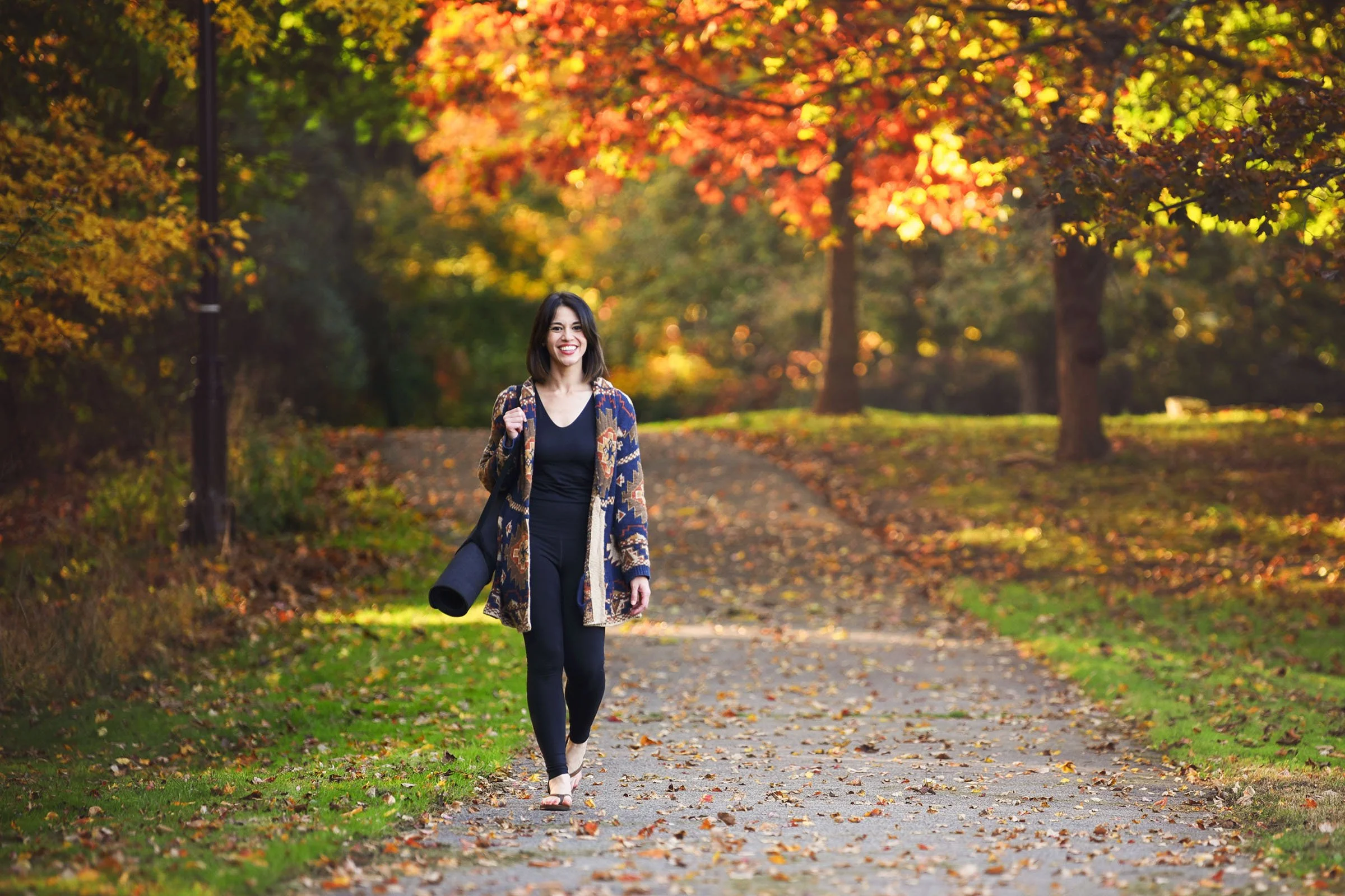 A woman with shoulder-length dark hair, wearing a patterned coat and black clothing, walking on a leaf-covered path in a park during autumn with vibrant orange, yellow, and red foliage in the background.