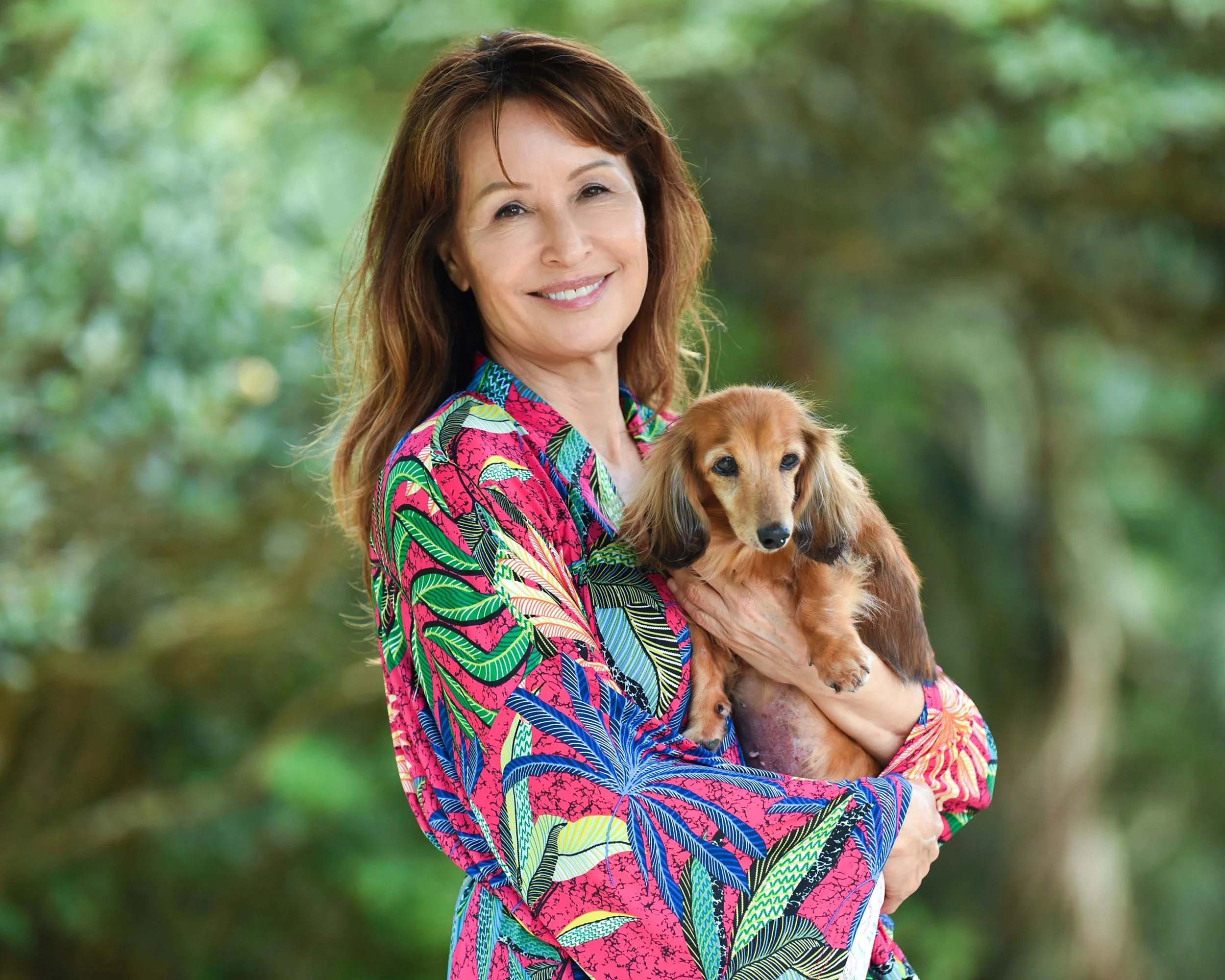 A middle-aged woman with brown hair and a bright smile, wearing a colorful patterned shirt, is holding a small, long-haired dachshund puppy outdoors with a blurred green background.