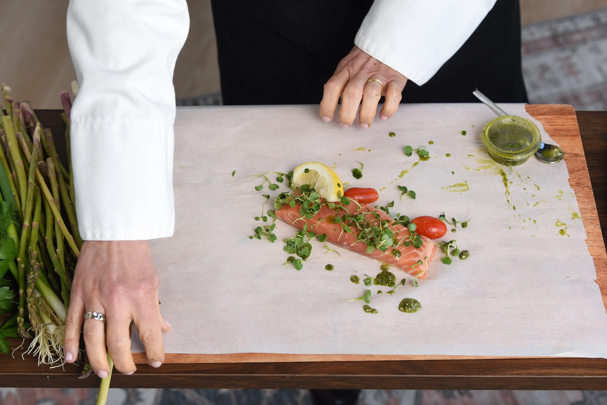 A person preparing a dish with fresh salmon, lemon slices, cherry tomatoes, herbs, and green sauce on a wooden cutting board, with asparagus on the side.