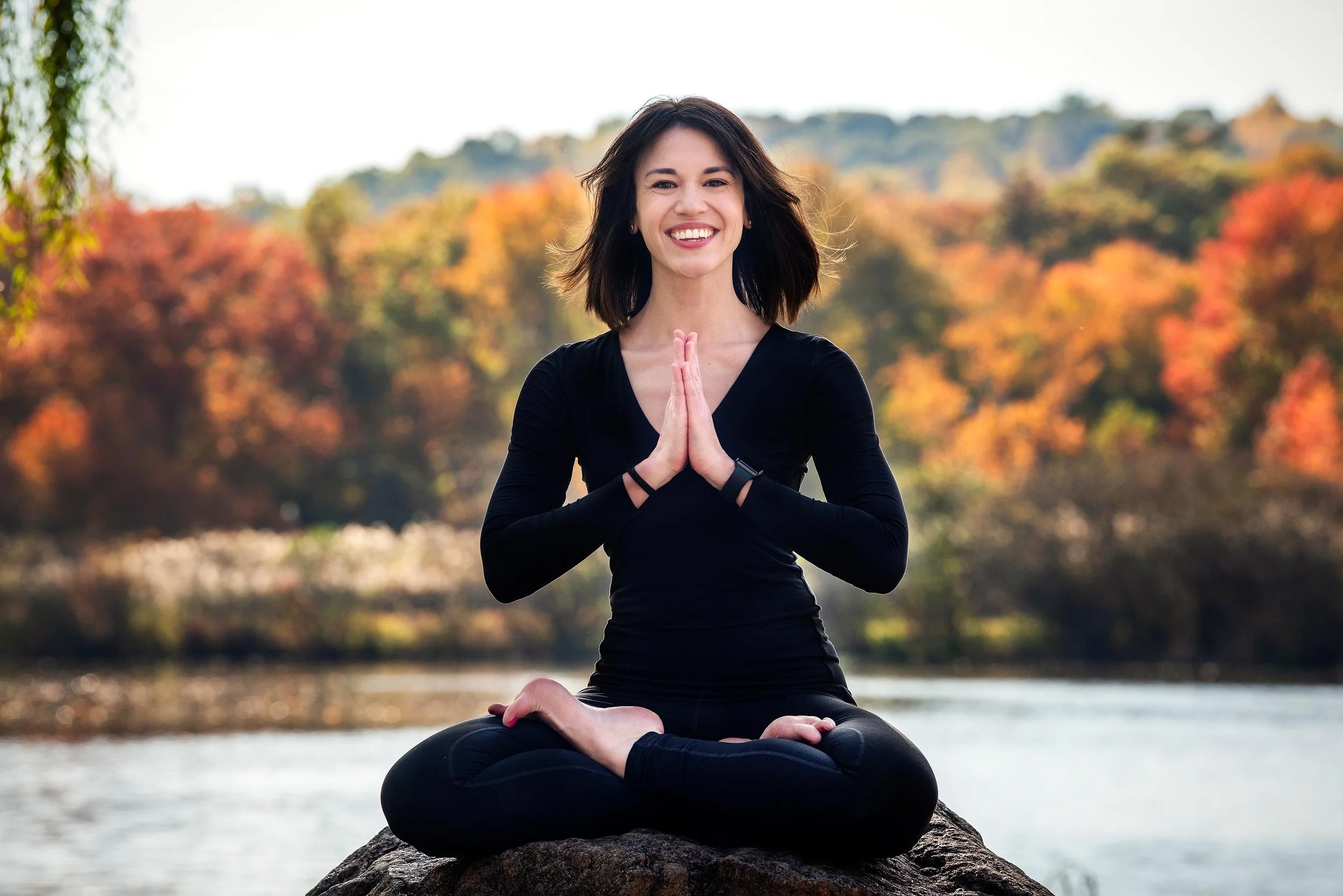 A woman practicing yoga outdoors near a lake during autumn with colorful trees in the background.