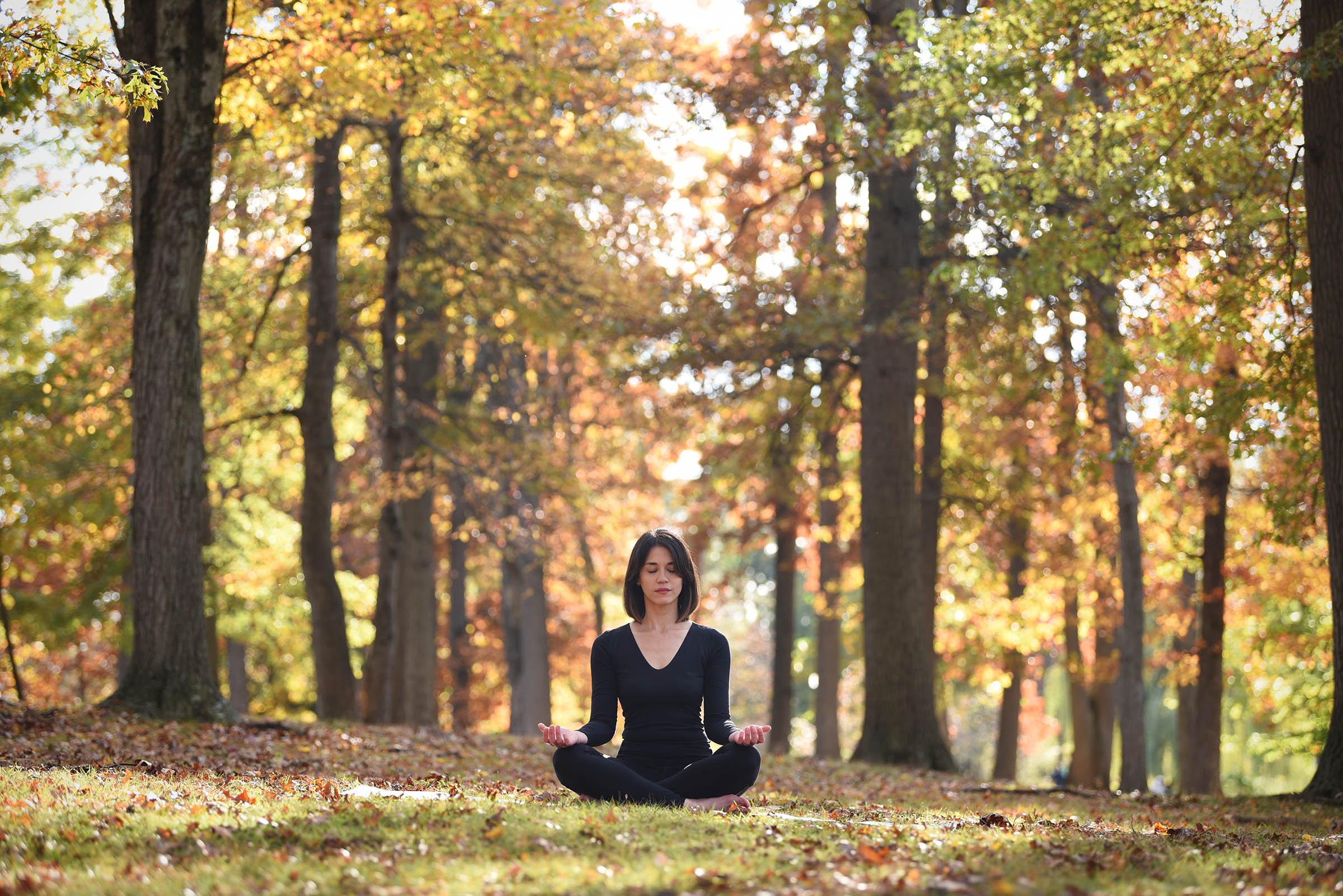 A woman practicing meditation outdoors in a park surrounded by autumn trees with colorful leaves.