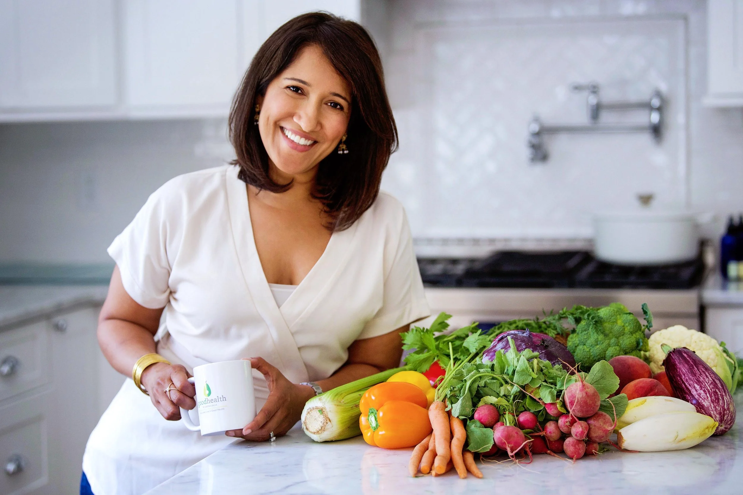 A woman with shoulder-length brown hair, wearing a white top and earrings, smiling while holding a coffee mug, stands in front of a table filled with various fresh vegetables including carrots, bell peppers, radishes, cauliflower, and broccoli, in a 