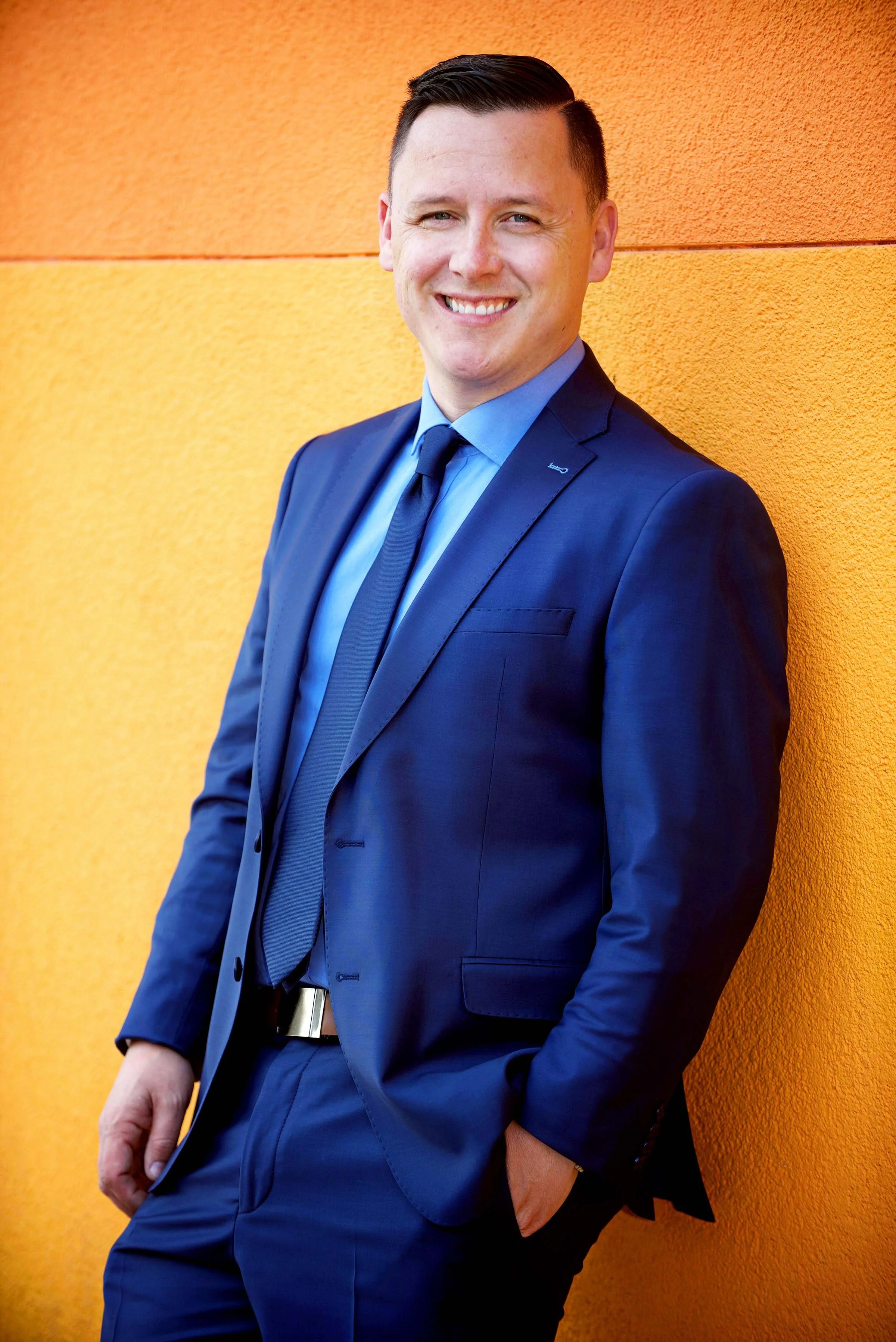 Portrait of a smiling man in a blue suit and tie standing against an orange textured wall.