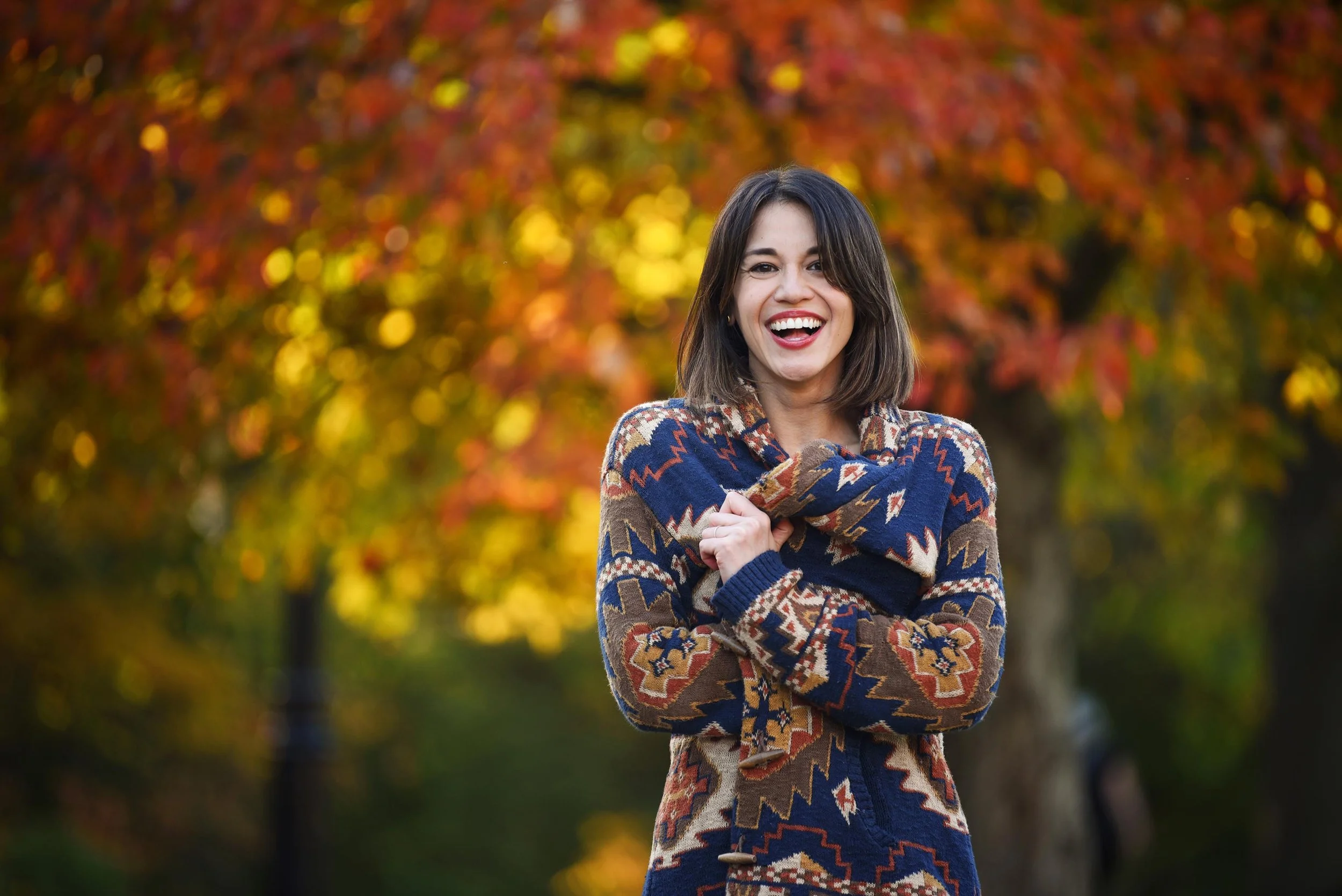 A woman standing outdoors in front of trees with colorful autumn leaves, smiling and wearing a patterned sweater.