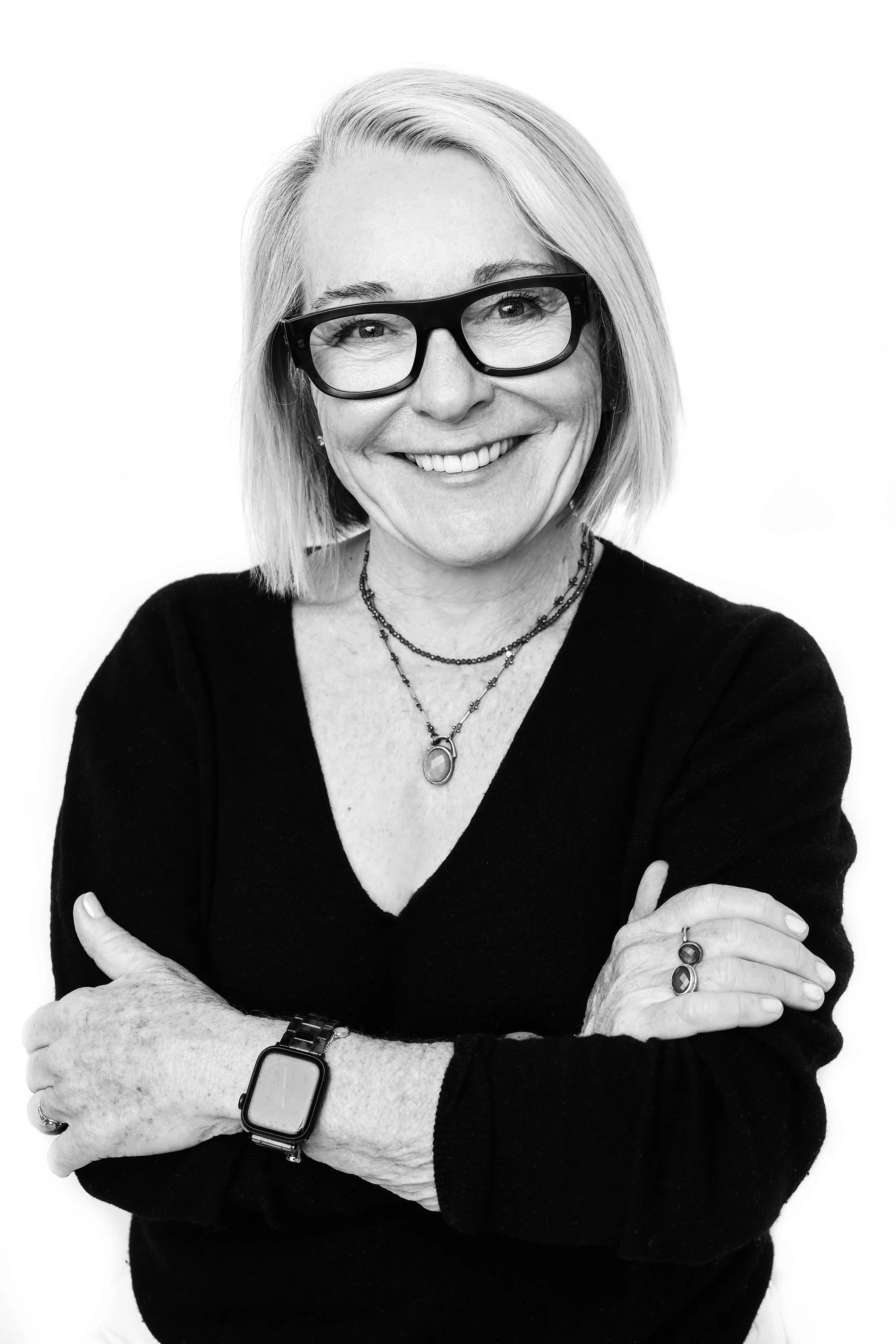 Black and white portrait of a smiling woman with shoulder-length blonde hair, wearing glasses, layered necklaces, a black top, a smartwatch, and jewelry on her fingers, with arms crossed in front of a plain white background.