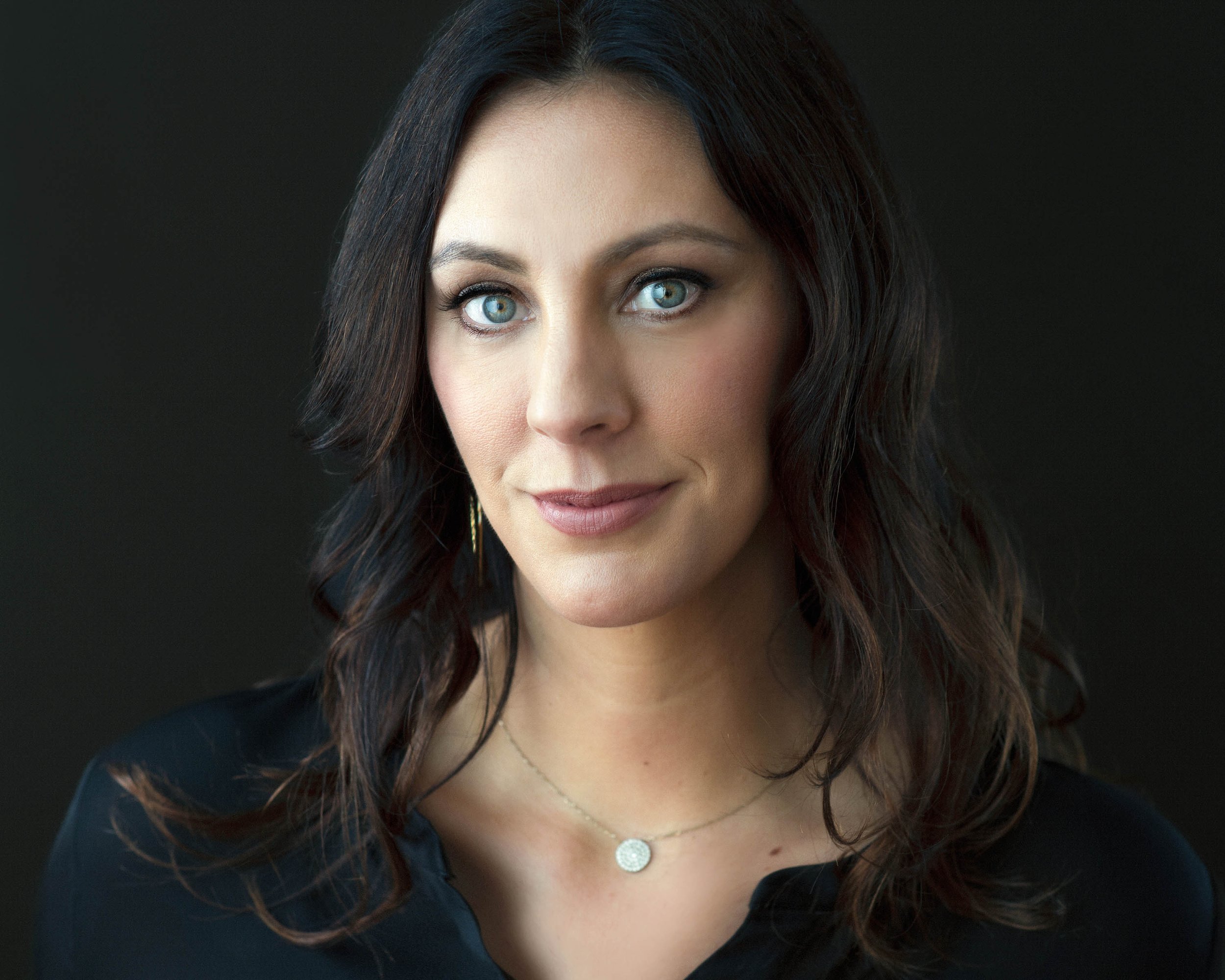 A woman with long dark hair and blue eyes looking at the camera, wearing a black top, a delicate necklace, and a small circular pendant, against a dark background.