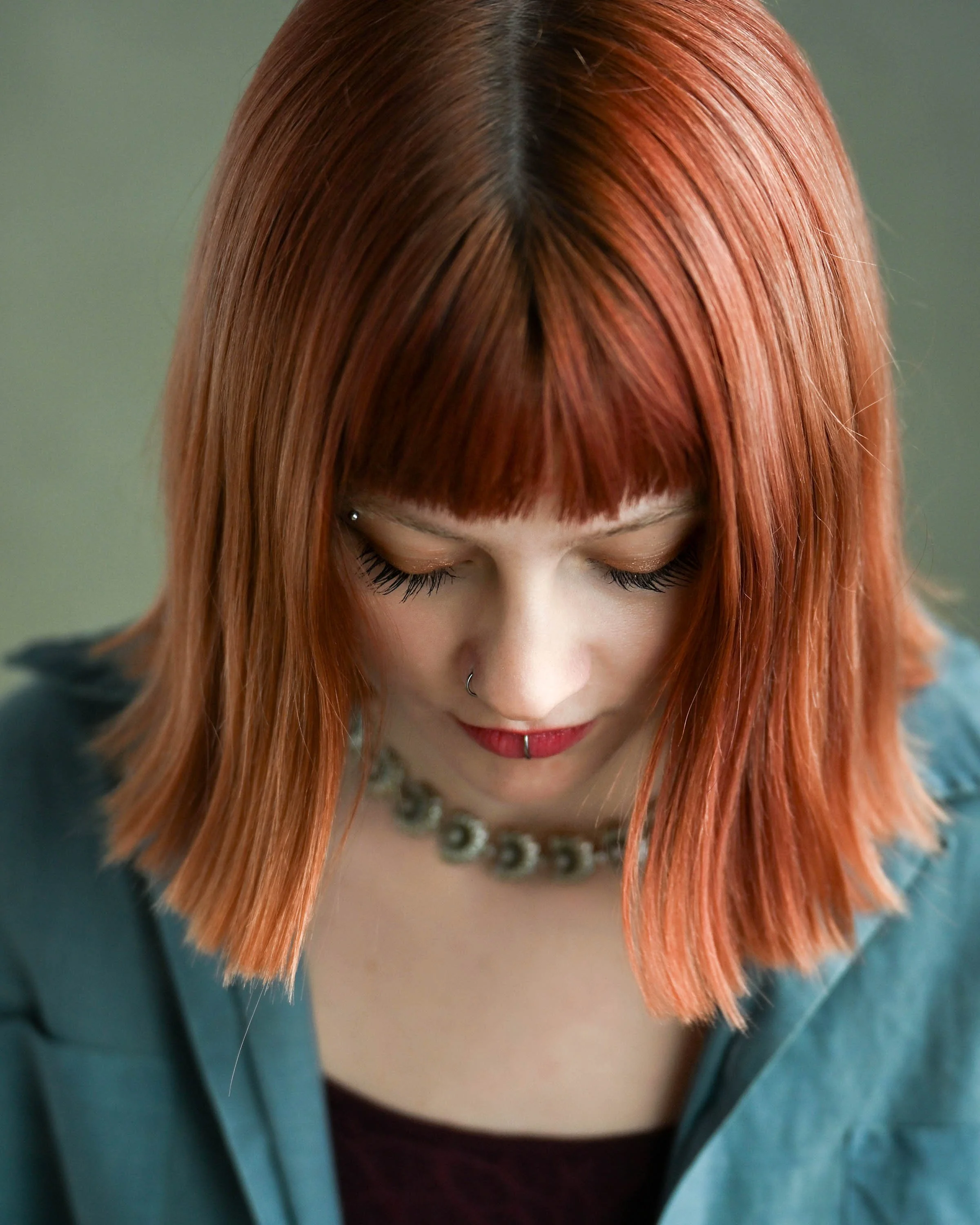Top view of a woman with vibrant orange hair, wearing a pearl necklace, with eyes closed and a nose piercing, dressed in a dark top with a teal blazer.