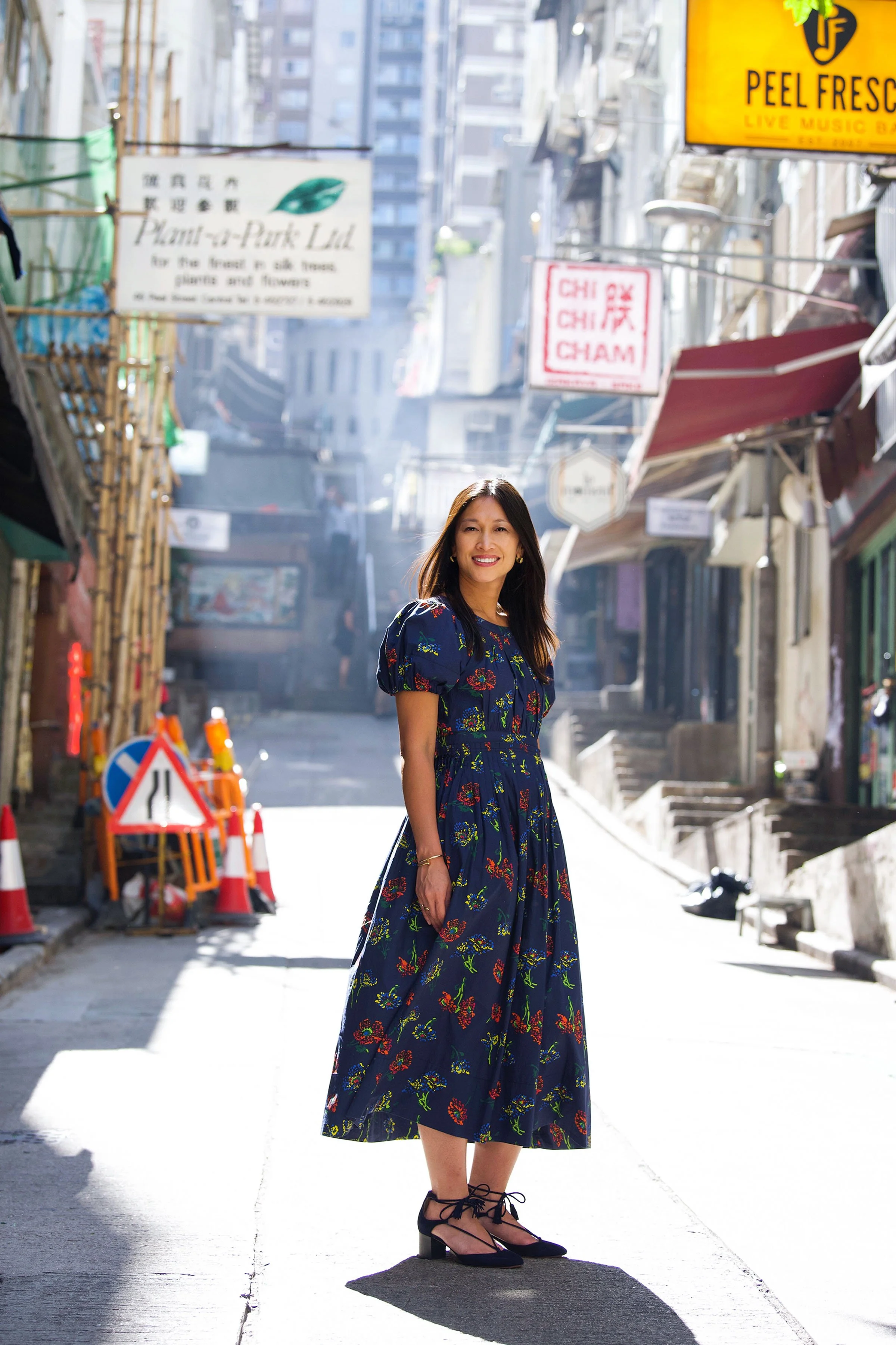 A woman in a long blue floral dress and black lace-up shoes standing on a city street with signs and scaffolding in the background.