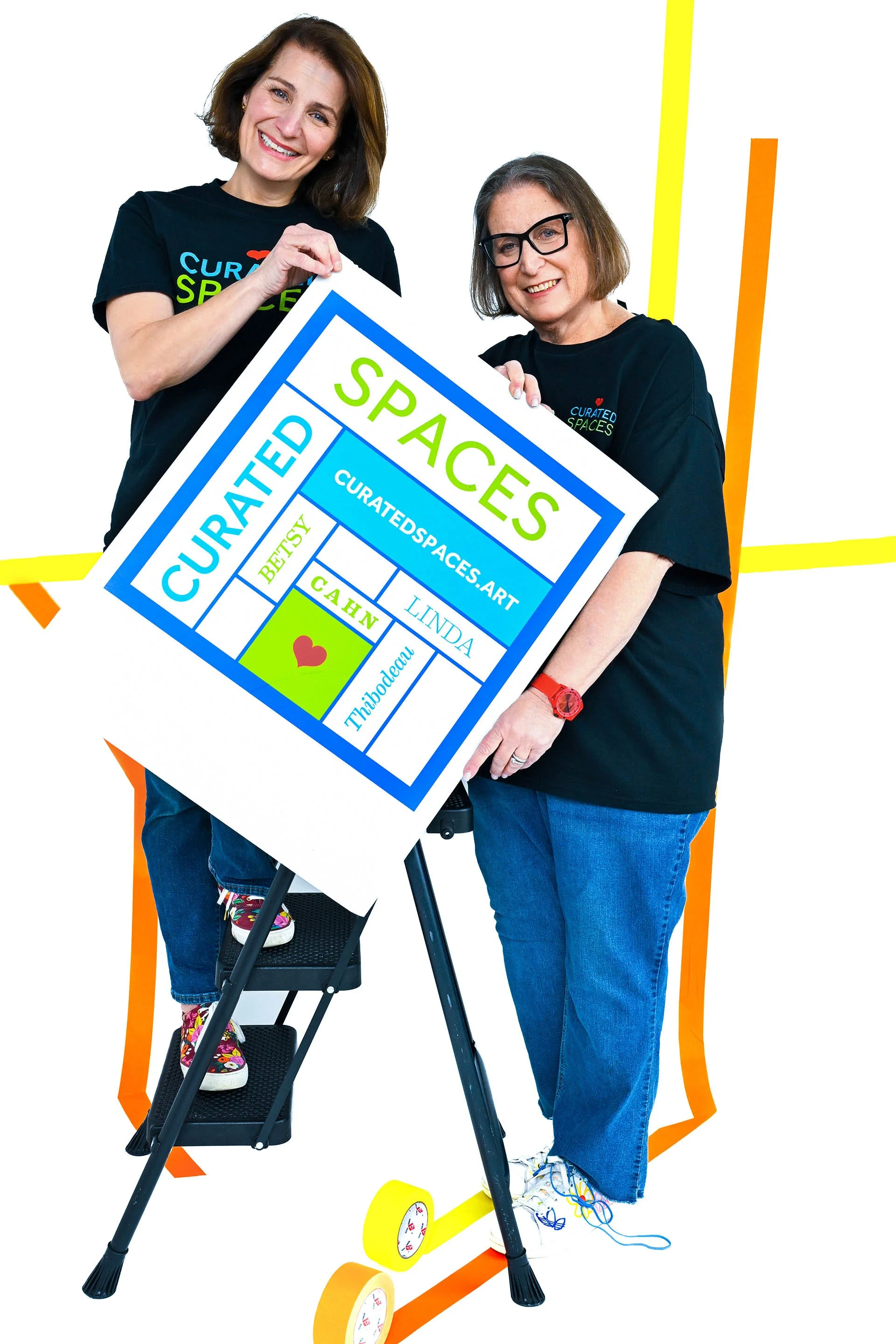 Two women holding a colorful poster that reads "Curated Spaces" with various smaller words and names, standing on a roller skateboard with orange and yellow accents. Both women are smiling; one is wearing glasses and the other is wearing sneakers wit