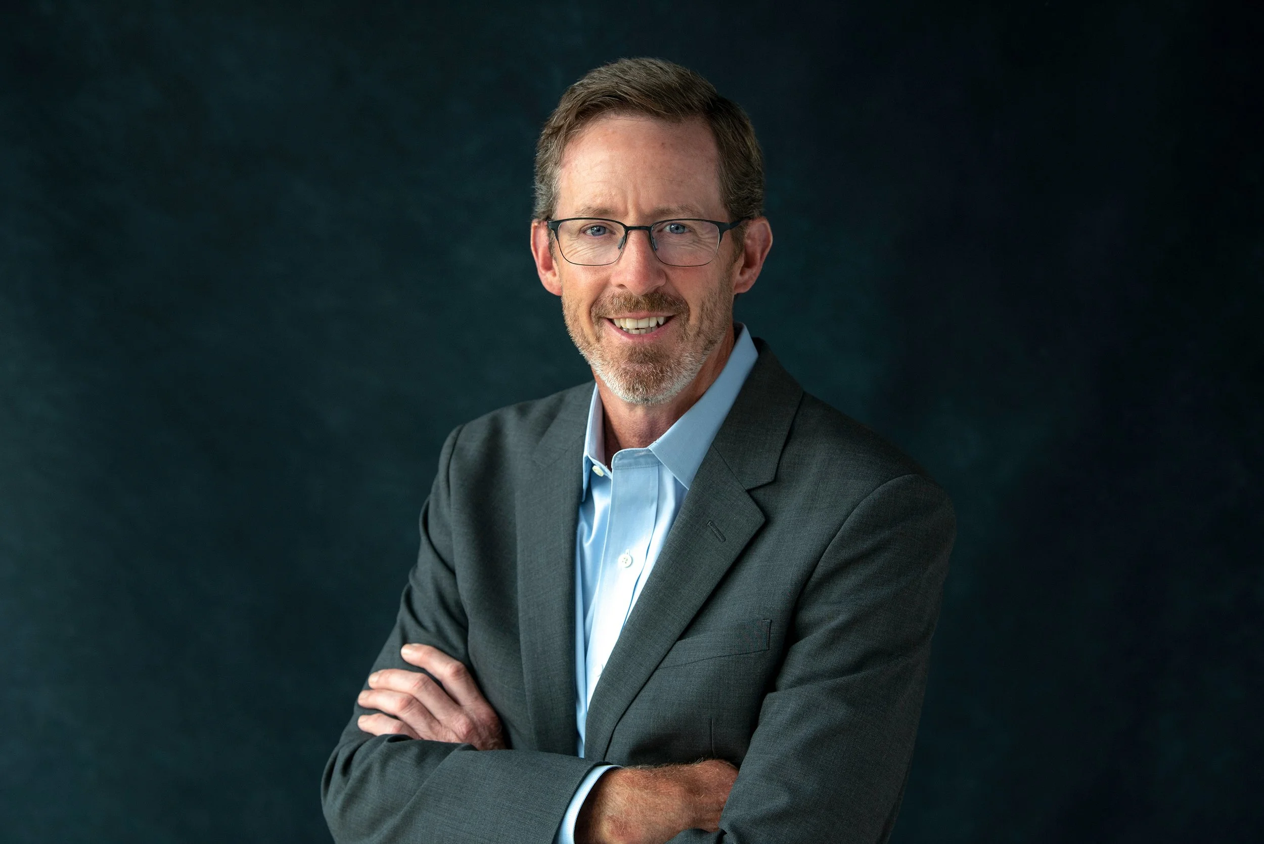 A middle-aged man wearing glasses and a gray suit smiling confidently with arms crossed in front of a dark background.