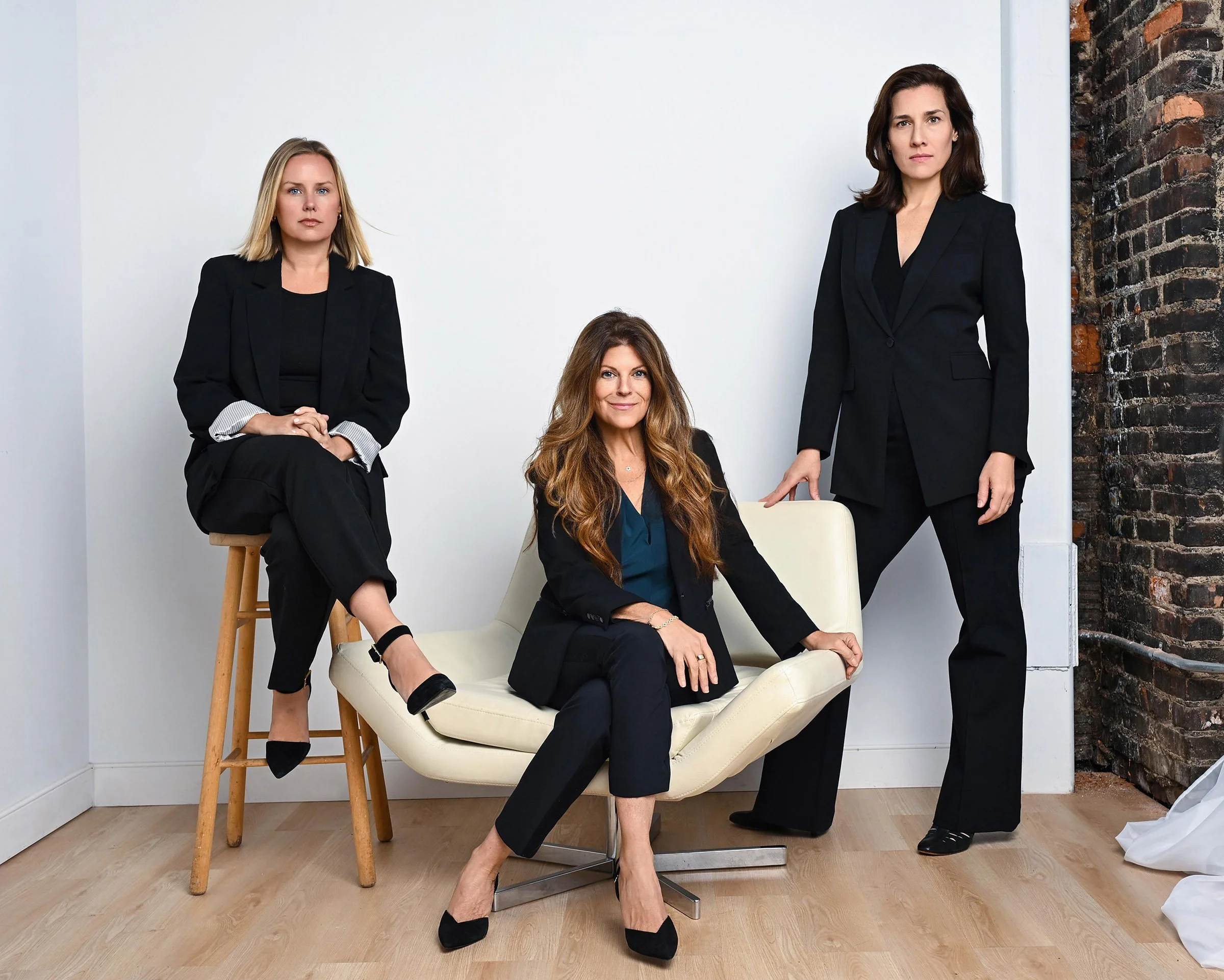 Four women dressed in black business attire posing in a modern minimalist indoor setting with a white wall and a brick wall. One woman is sitting on a white chair, another on a stool, and two standing.