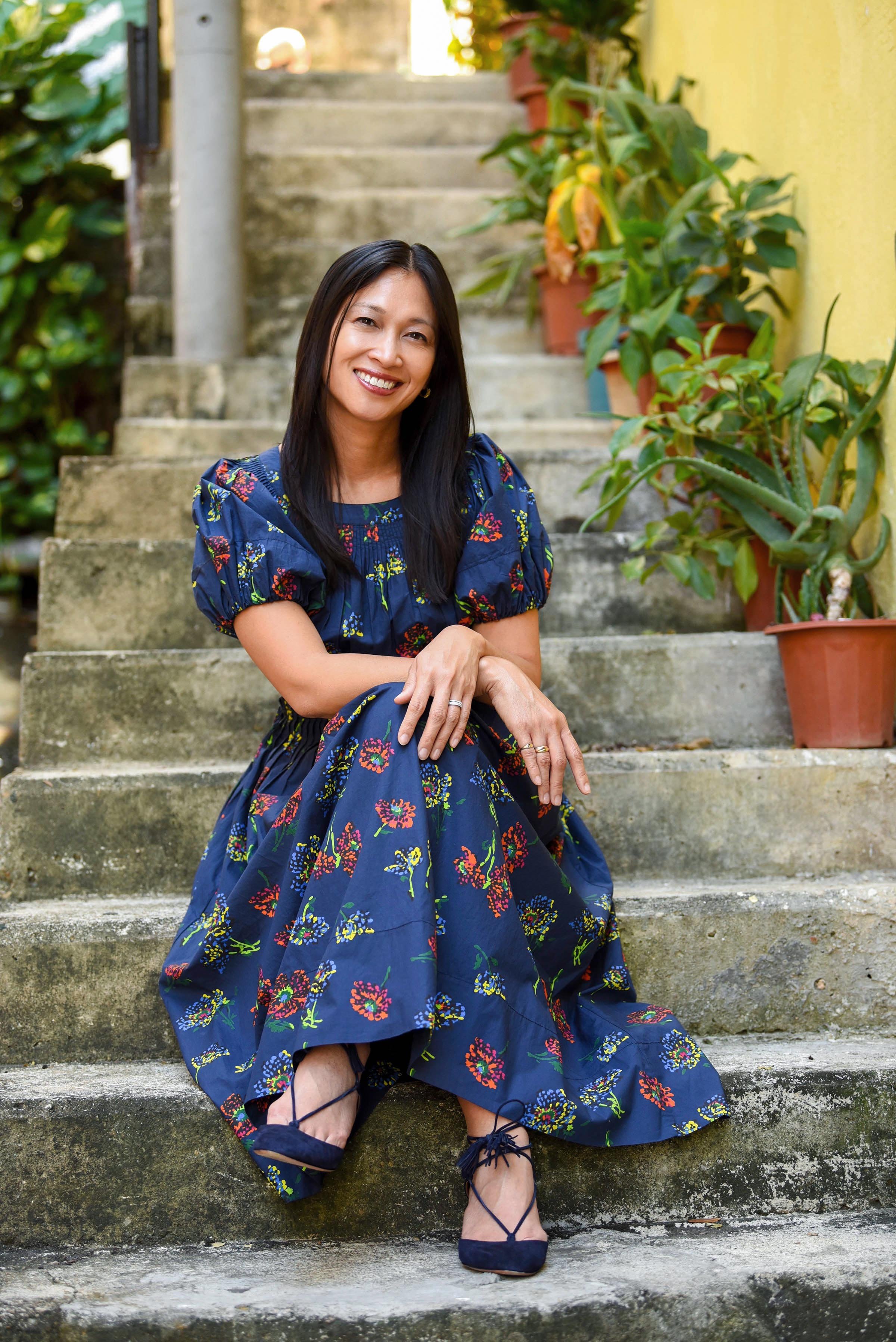 A woman with long dark hair and a bright smile sitting on concrete steps with potted plants and a yellow wall in the background, wearing a blue floral dress and blue shoes.