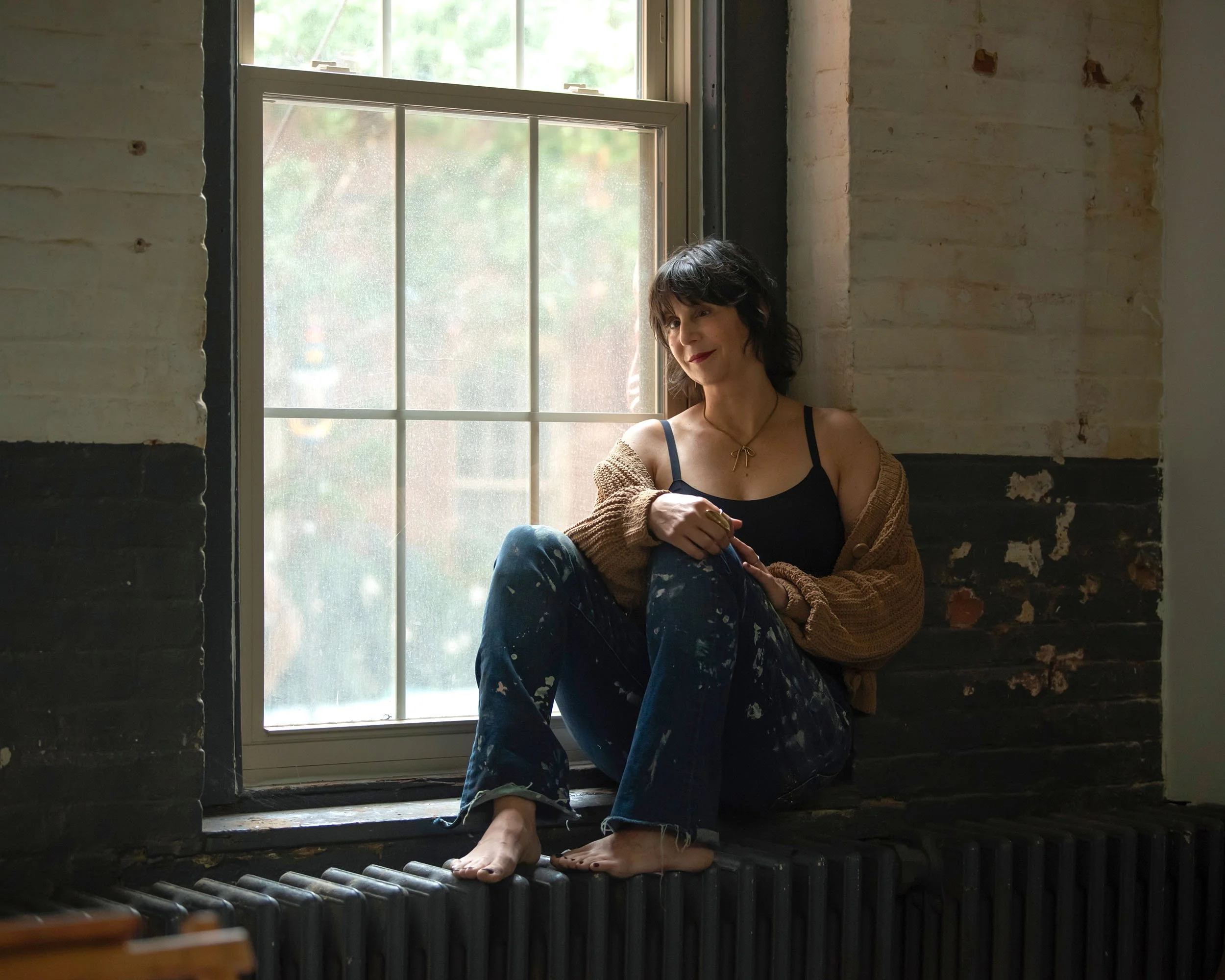 A woman with dark curly hair, wearing a black tank top and a beige cardigan, sits by a window with a brick wall behind her. She is smiling and looking slightly to the right. The window lets in natural light, and the view outside appears green and lea