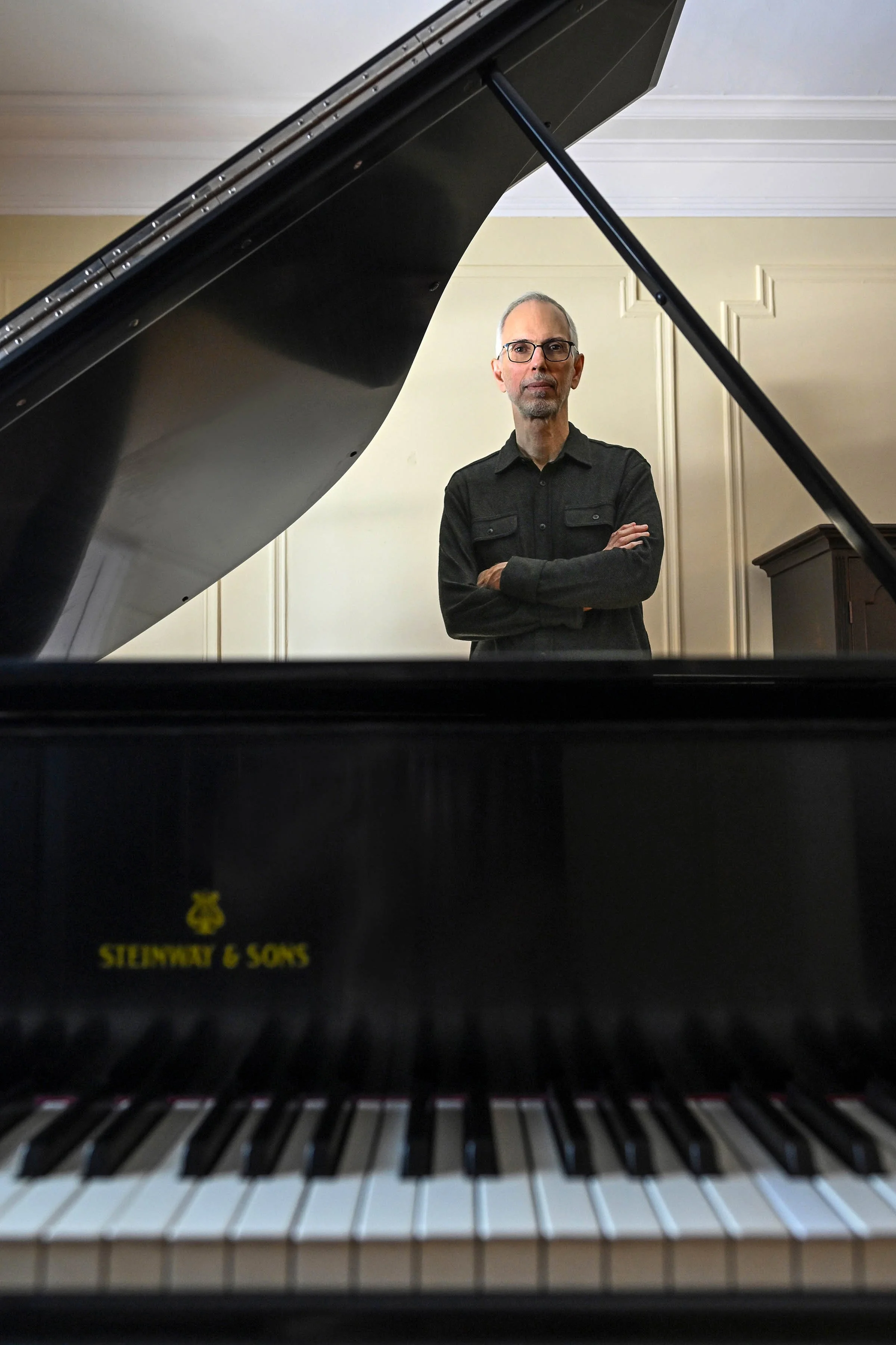 View from inside a grand piano looking toward the keyboard with a man standing behind it, crossing his arms, in a room with beige walls and paneled ceiling.