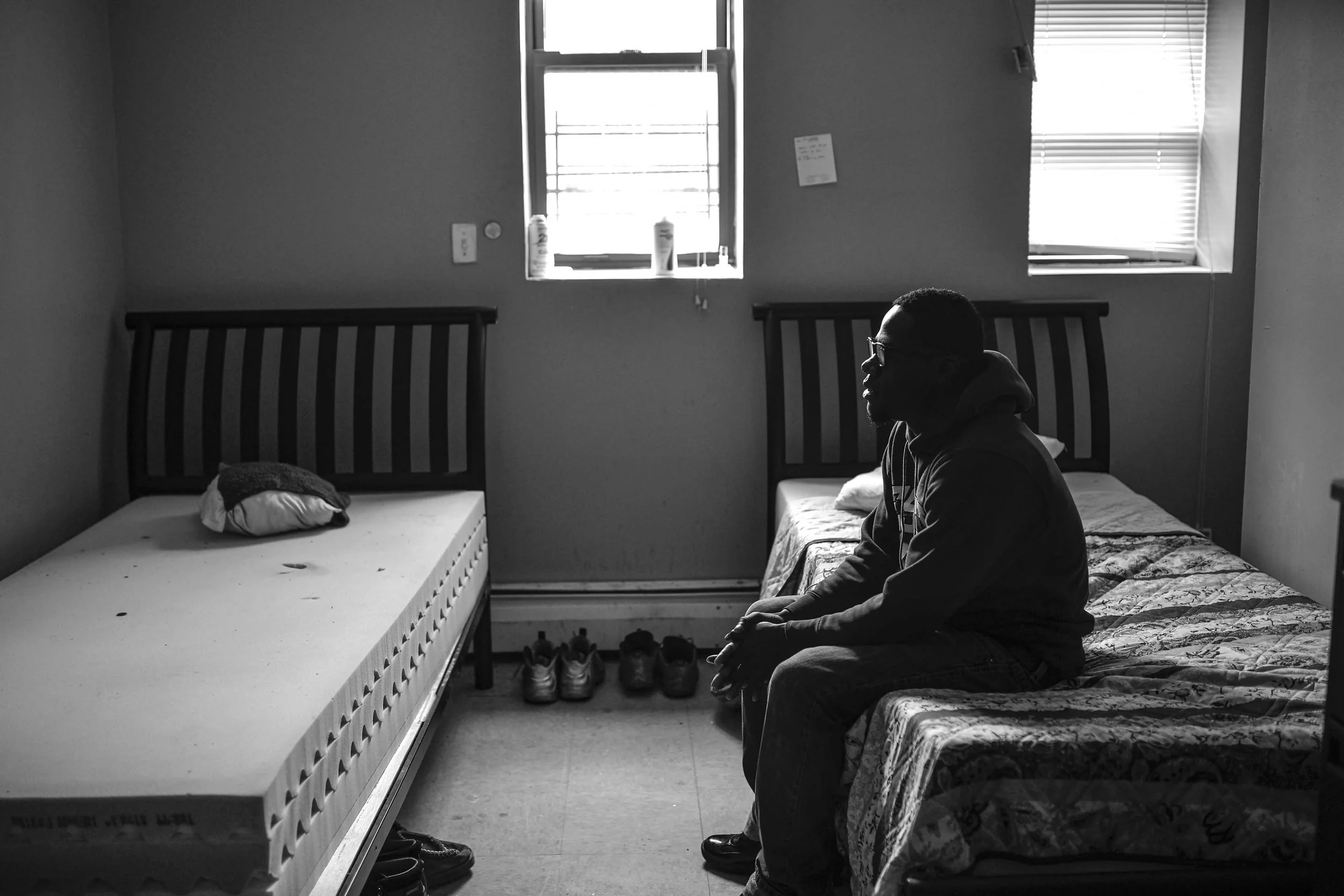 A grayscale photo of a person sitting on a bed in a small room, with two beds, two windows, and shoes on the floor.